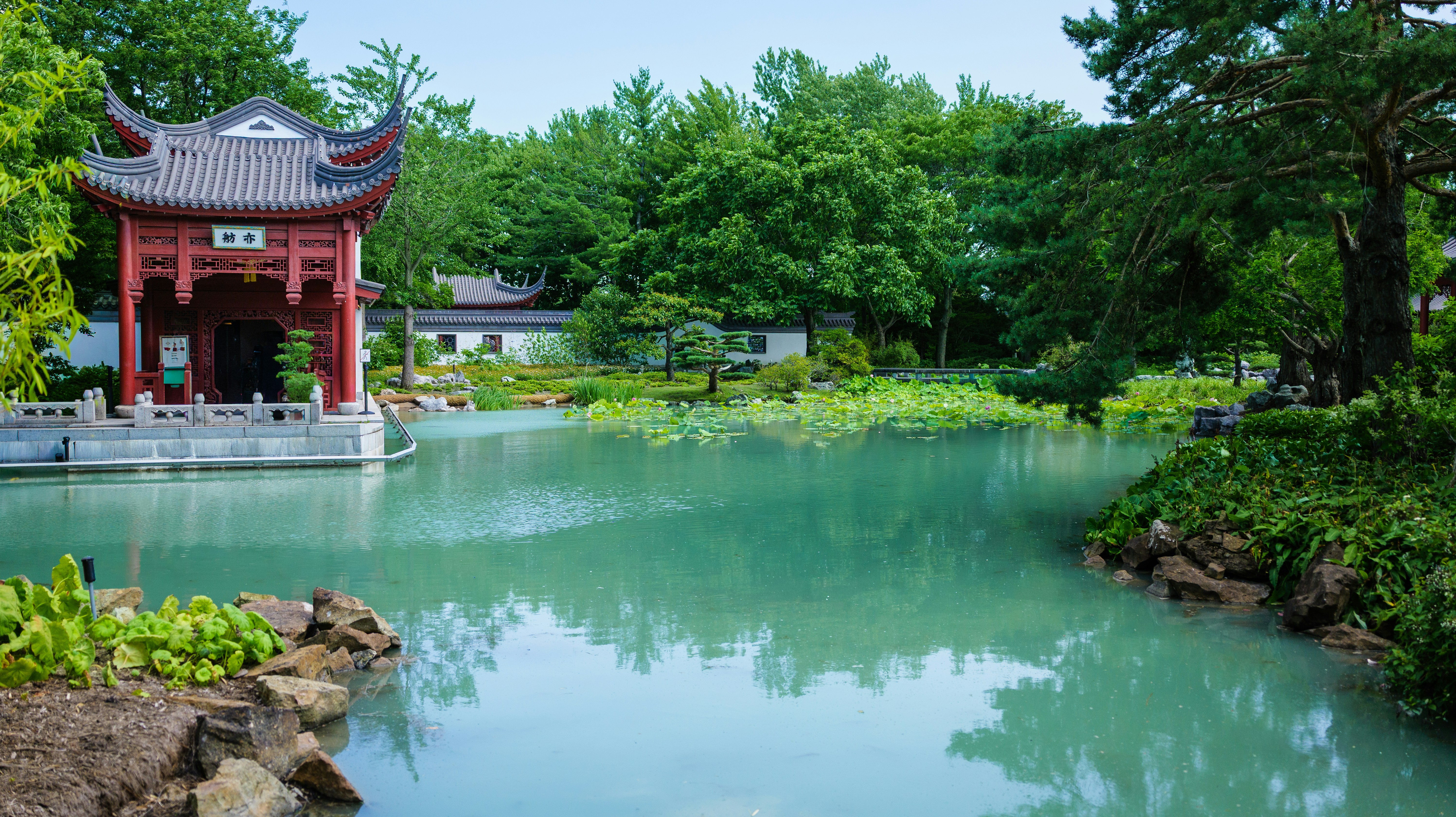 A traditional Chinese pavilion beside a turquoise pond in a lush garden