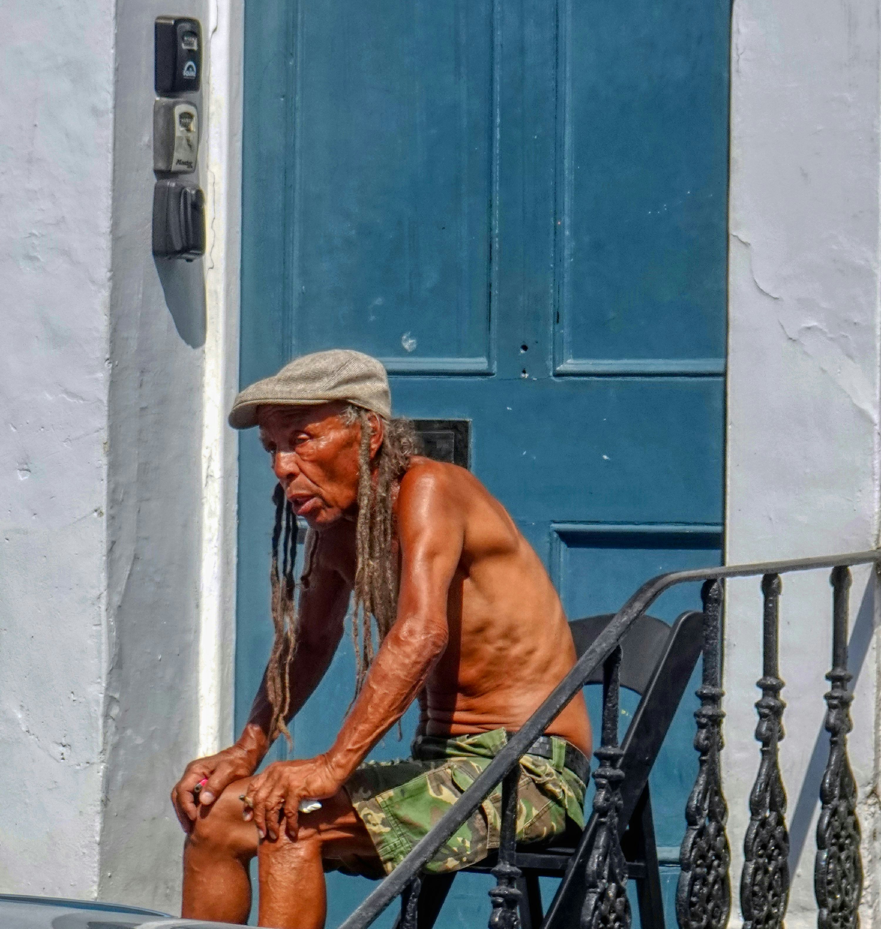 Un homme plus âgé aux longues dreadlocks, portant une casquette et un short camouflage, sur une chaise
