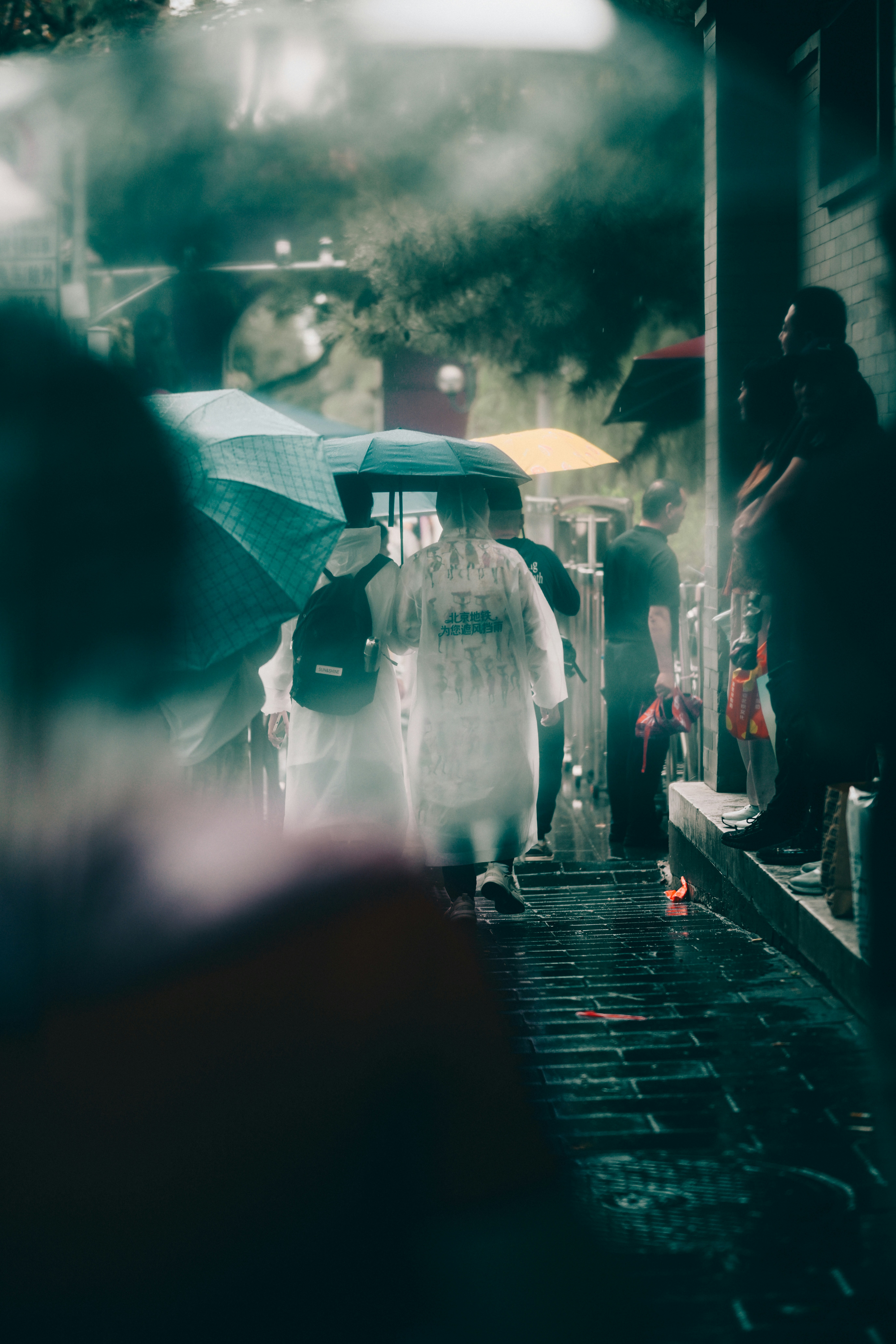 People in raincoats and umbrellas walking on a wet brick sidewalk in Beijing