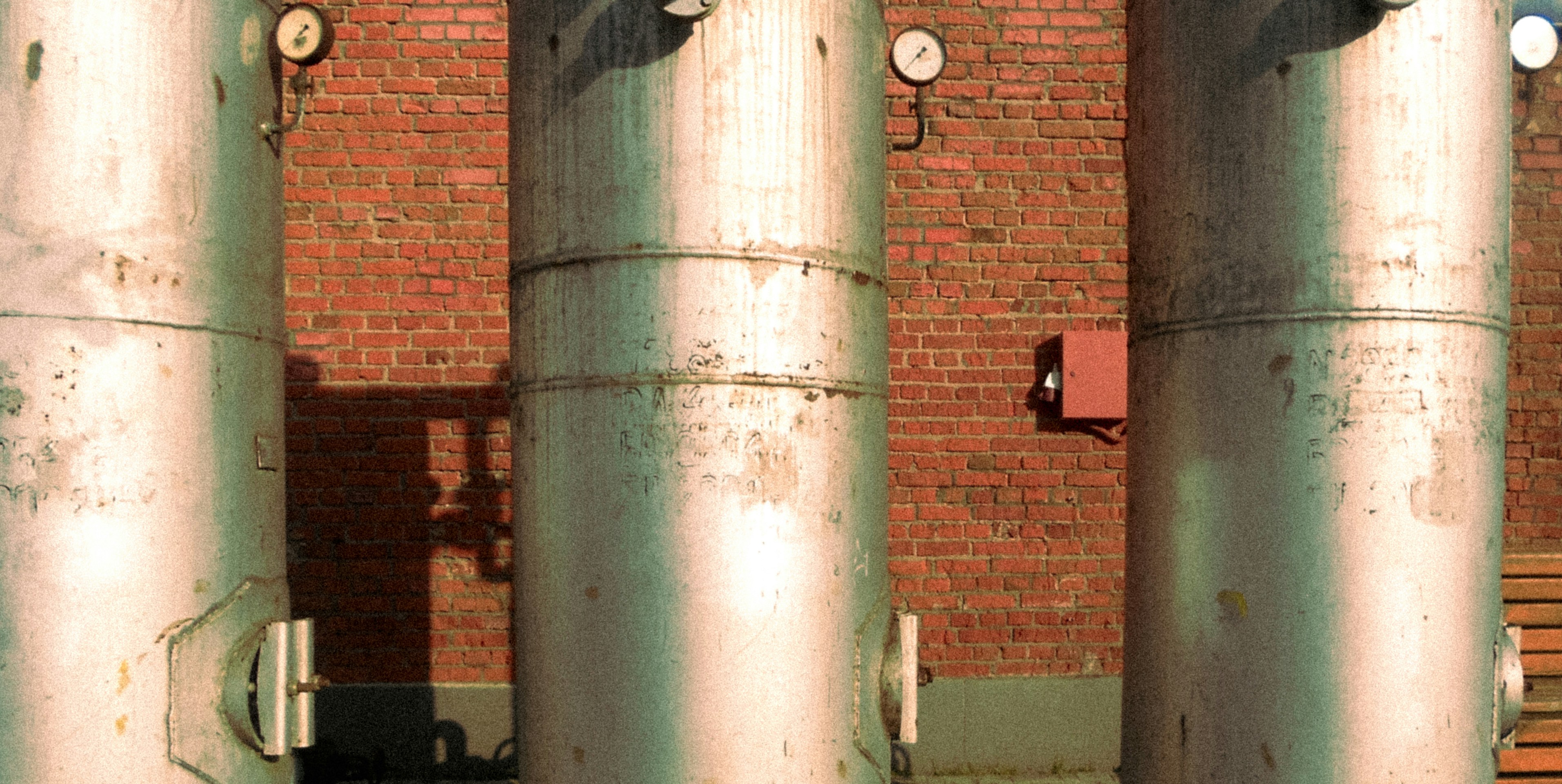 Three tall metal tanks with pressure gauges against a brick wall