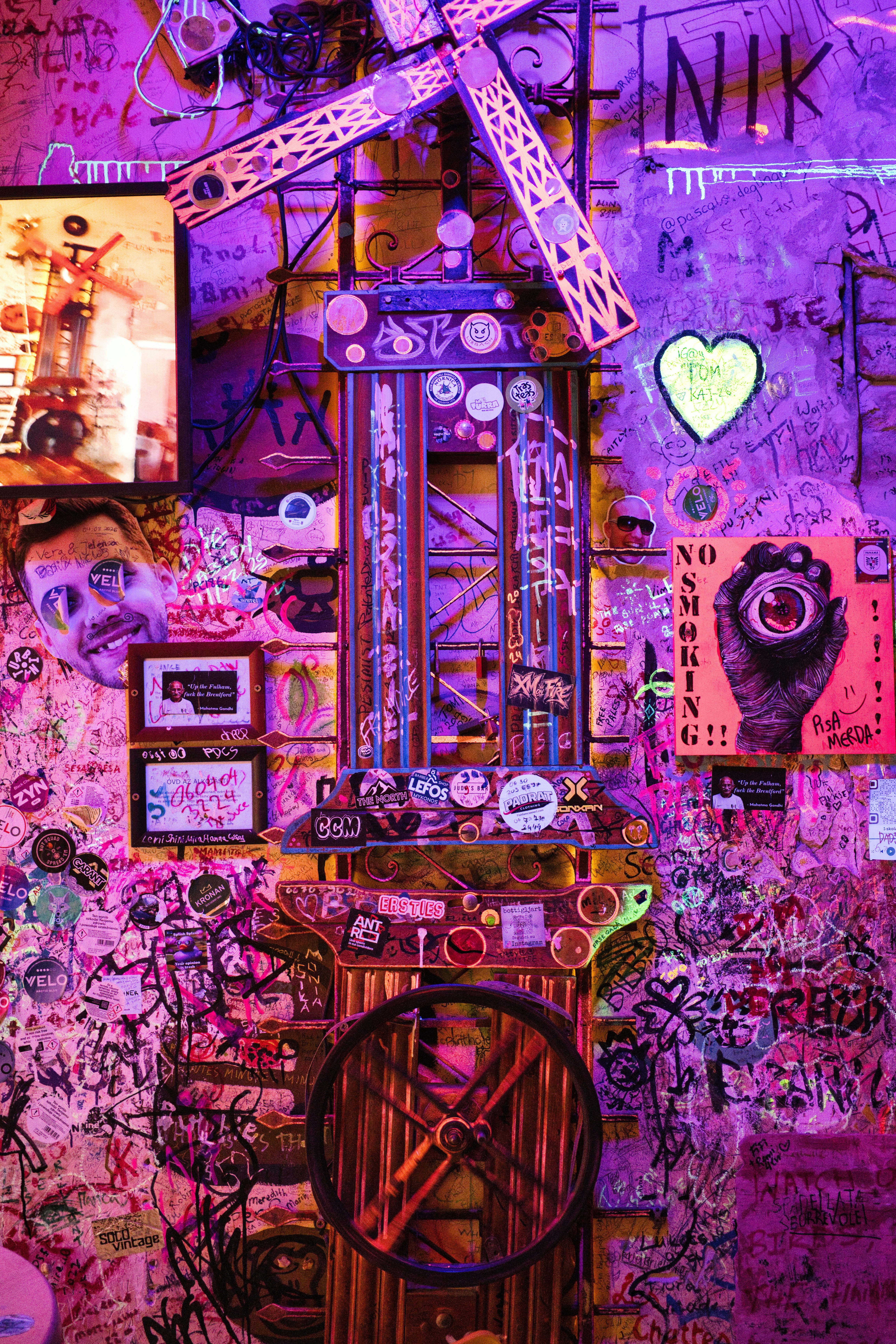 A metal windmill sculpture covered in stickers against a graffiti wall in purple light