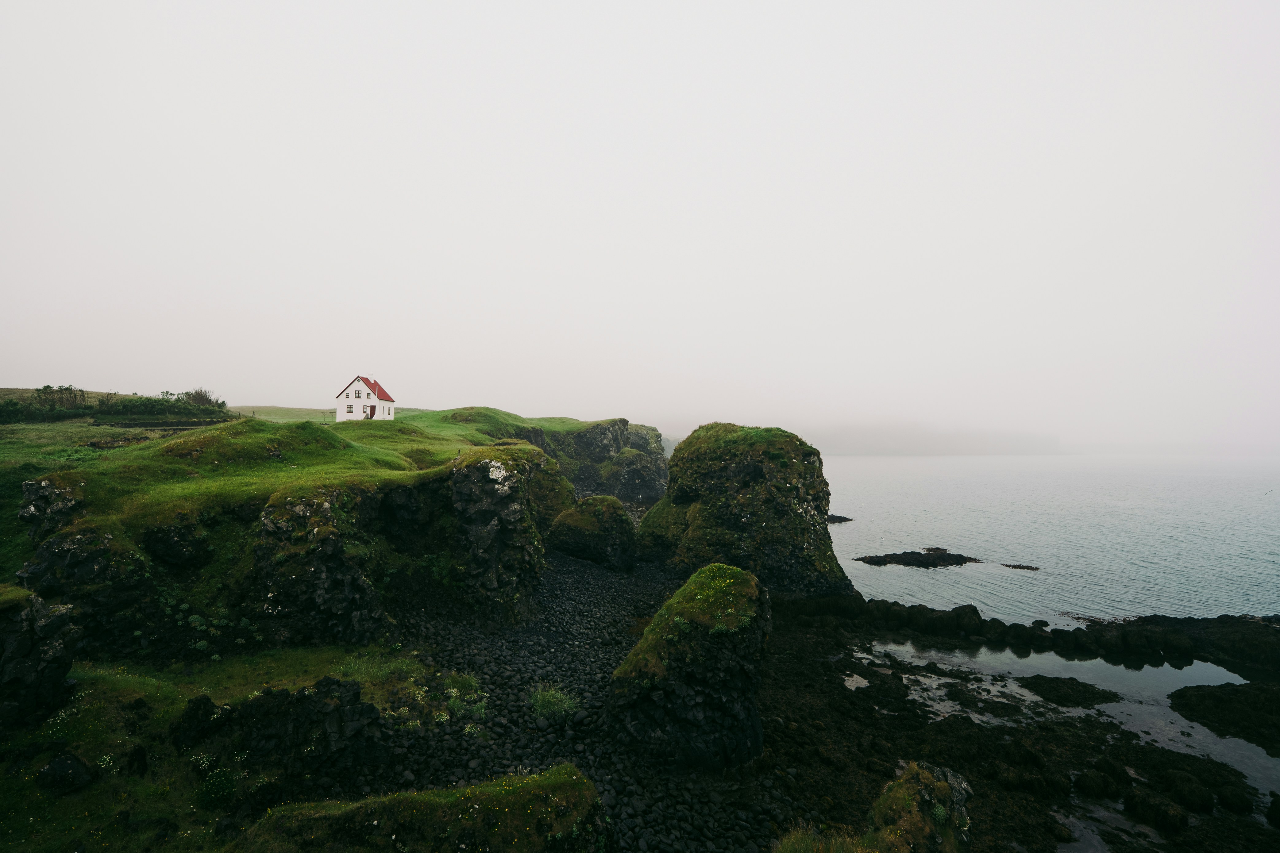 A small white house with a red roof on a grassy cliff above the ocean