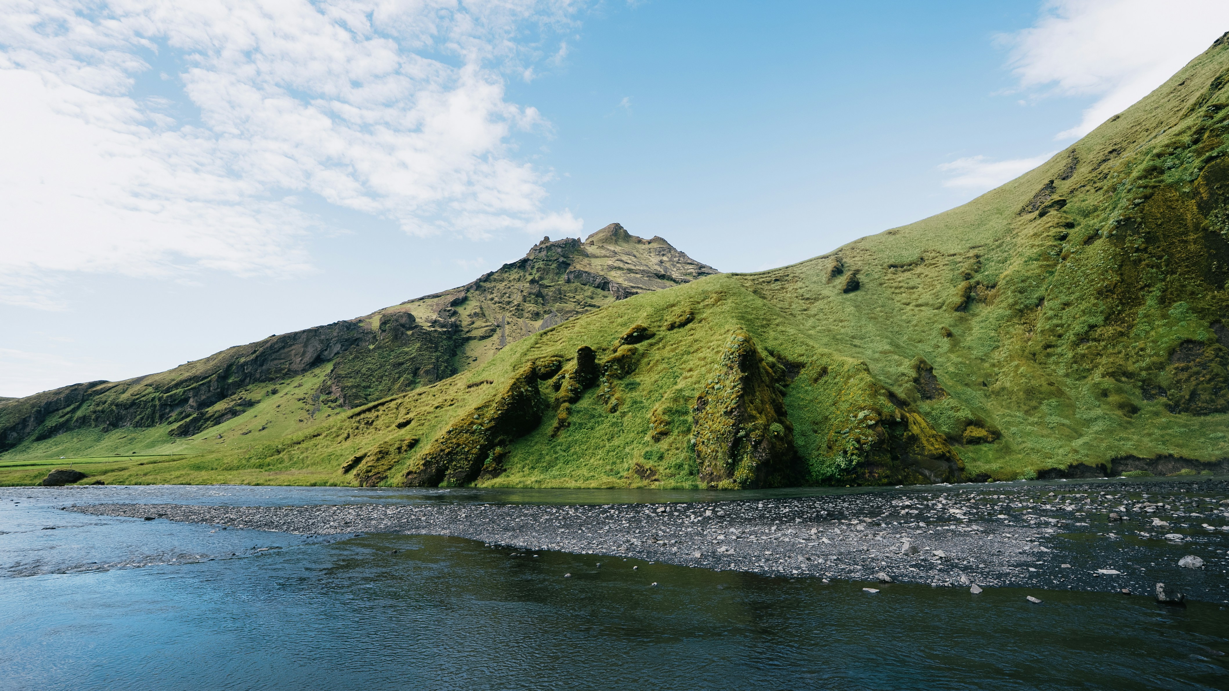 A river flowing past a steep green mountain slope under a blue sky