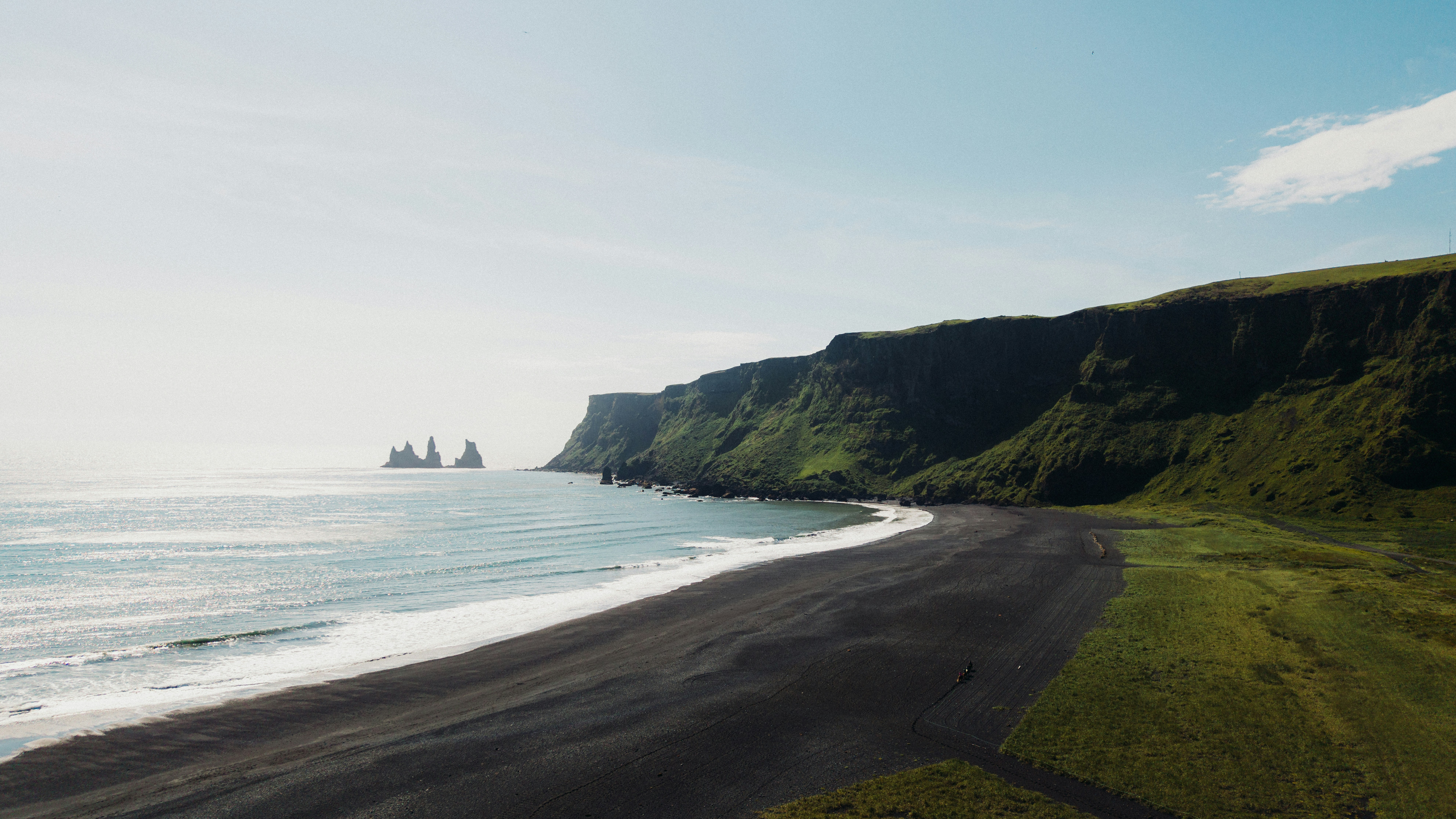 Black sand beach below green cliffs with sea stacks in the distance