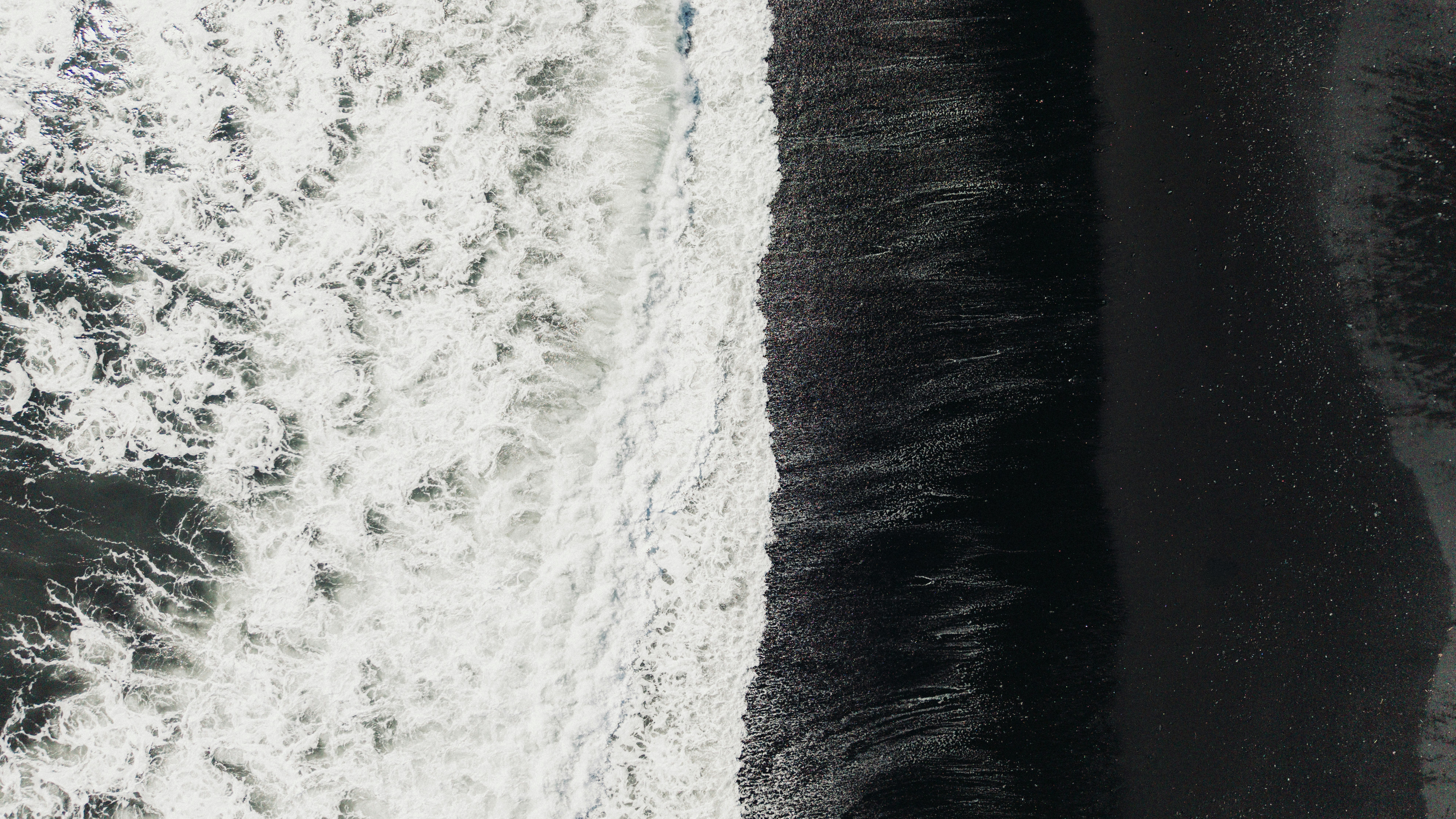 Aerial view of white ocean waves crashing onto a dark black sand beach