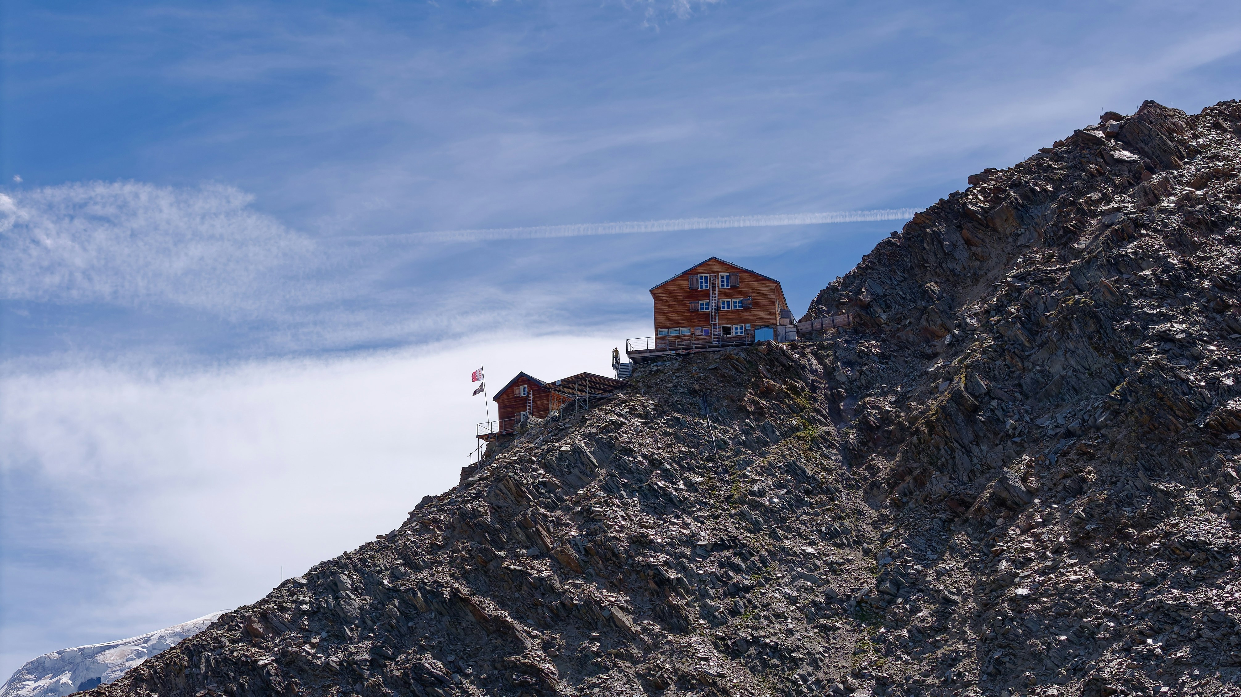 Two wooden mountain huts perched on a steep rocky ridge under a blue sky