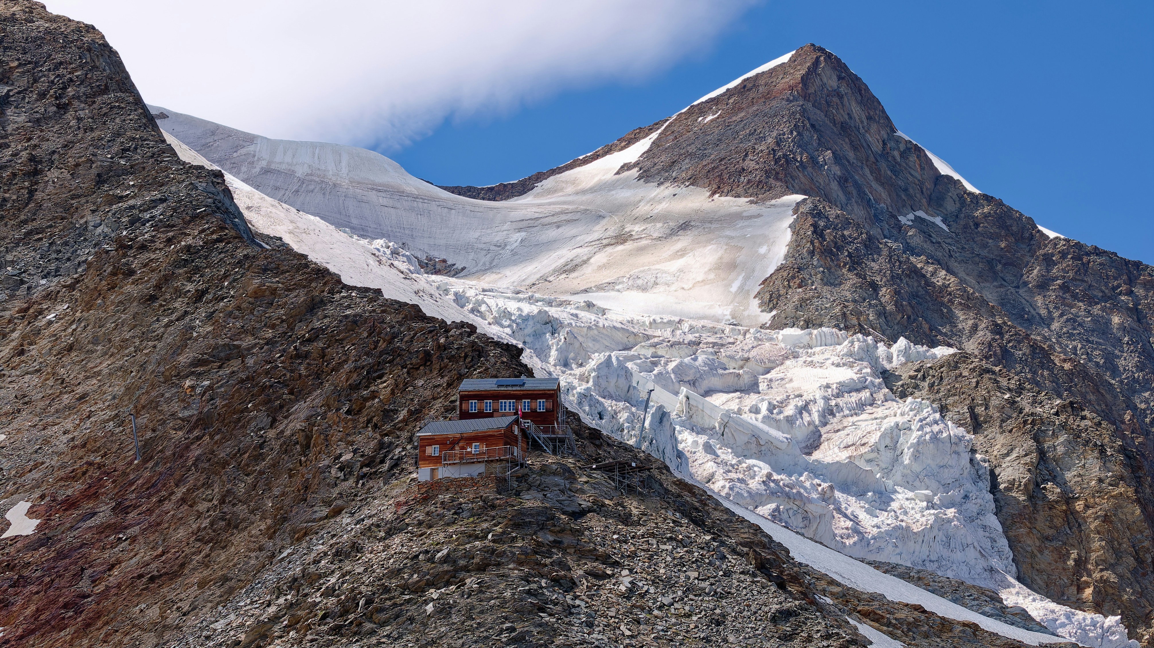 A wooden mountain hut perched on a rocky ridge below a massive glacier