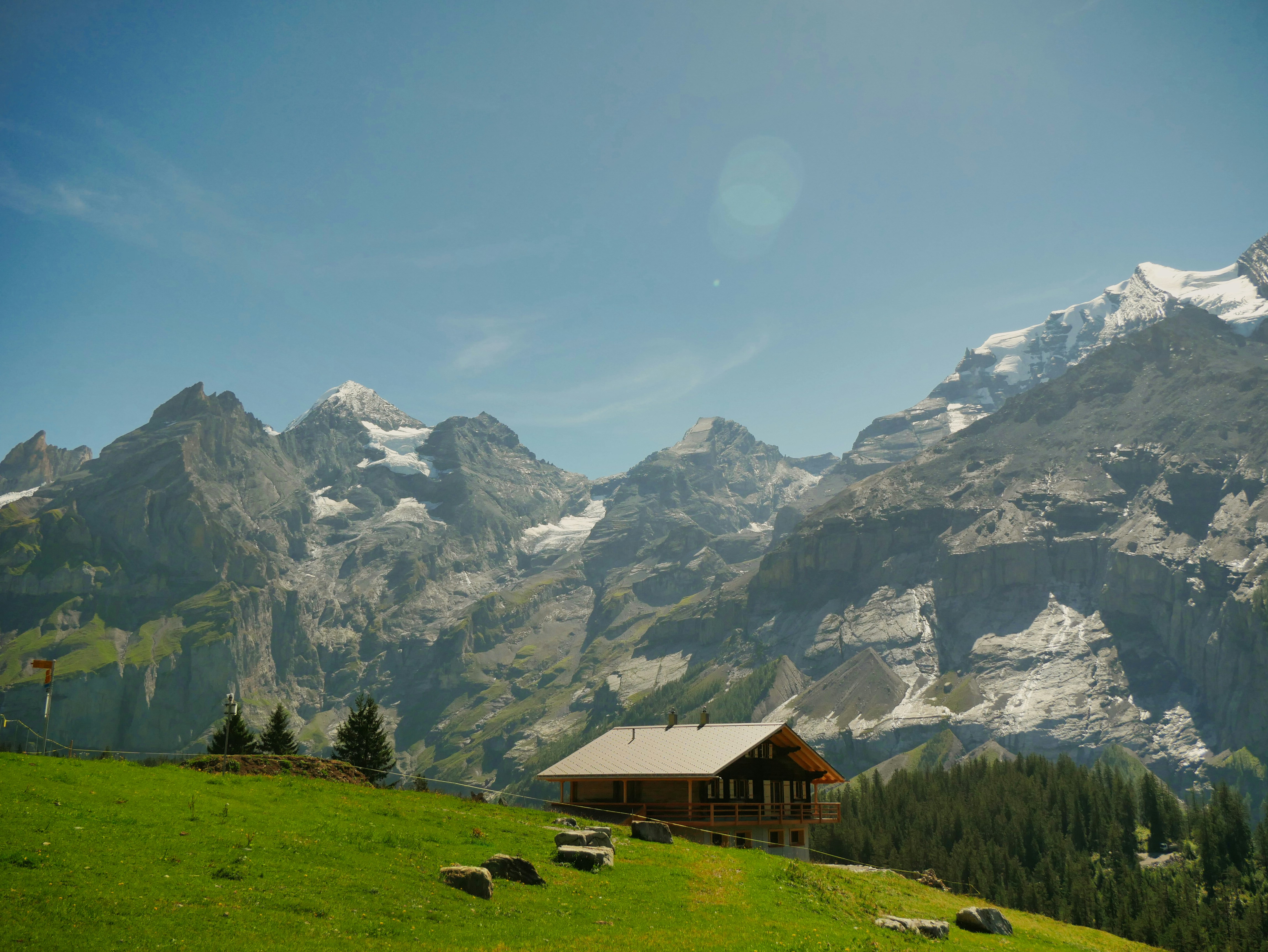 Wooden cabin on a green alpine meadow below jagged snow-capped mountain peaks