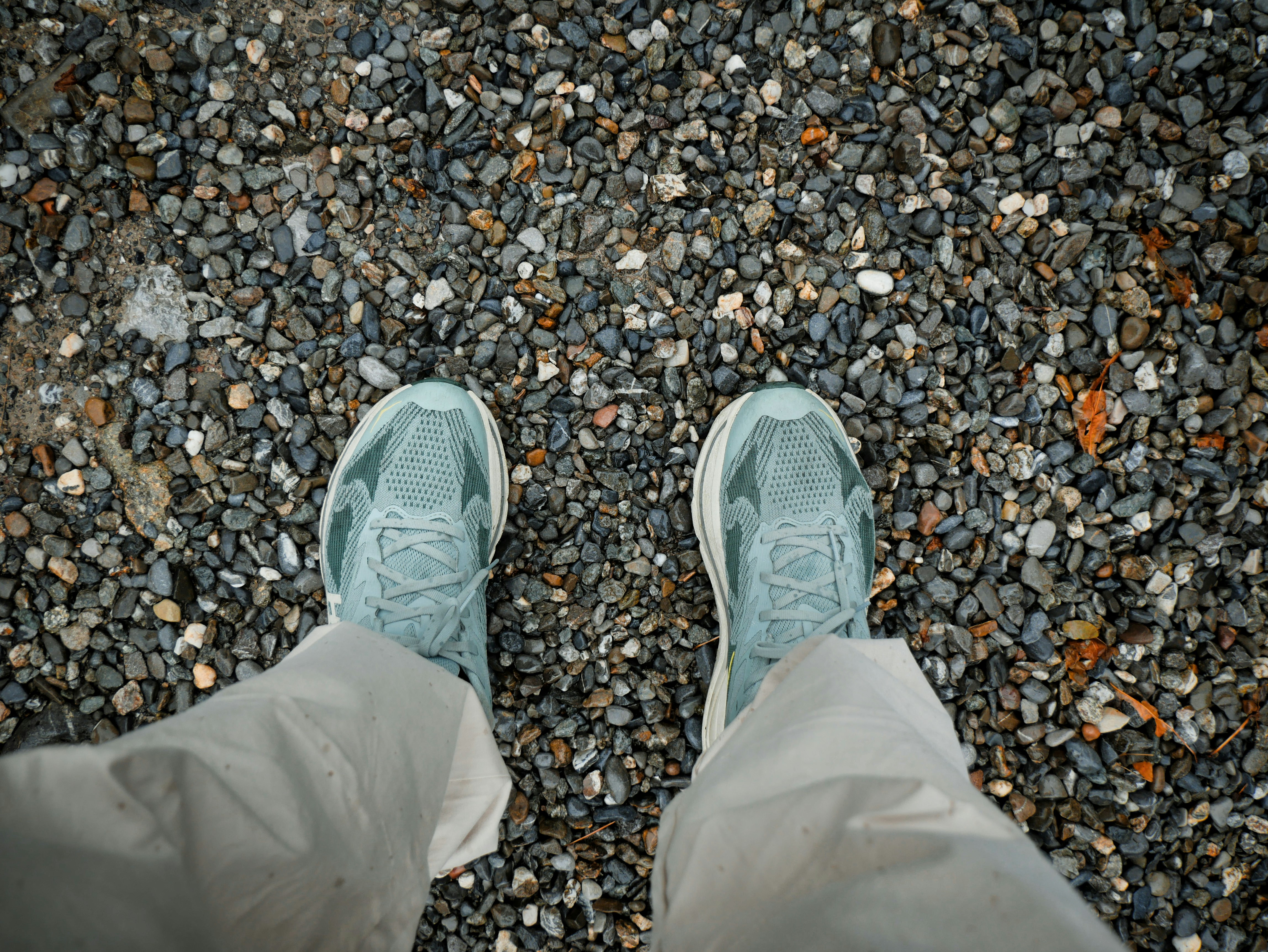 A person wearing light green sneakers and beige trousers standing on grey gravel