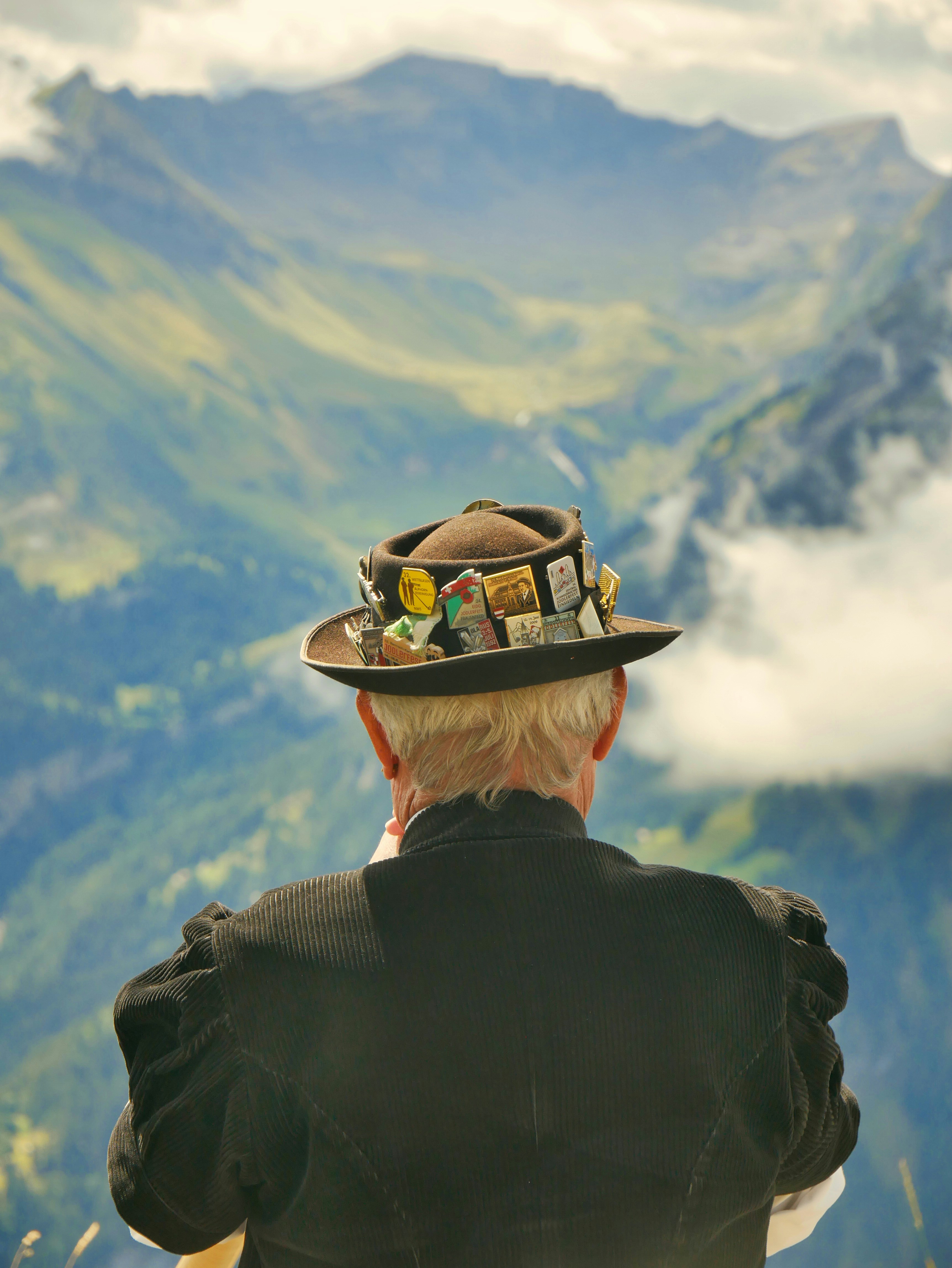 An older man seen from behind, his hat covered in pins, looking over a valley