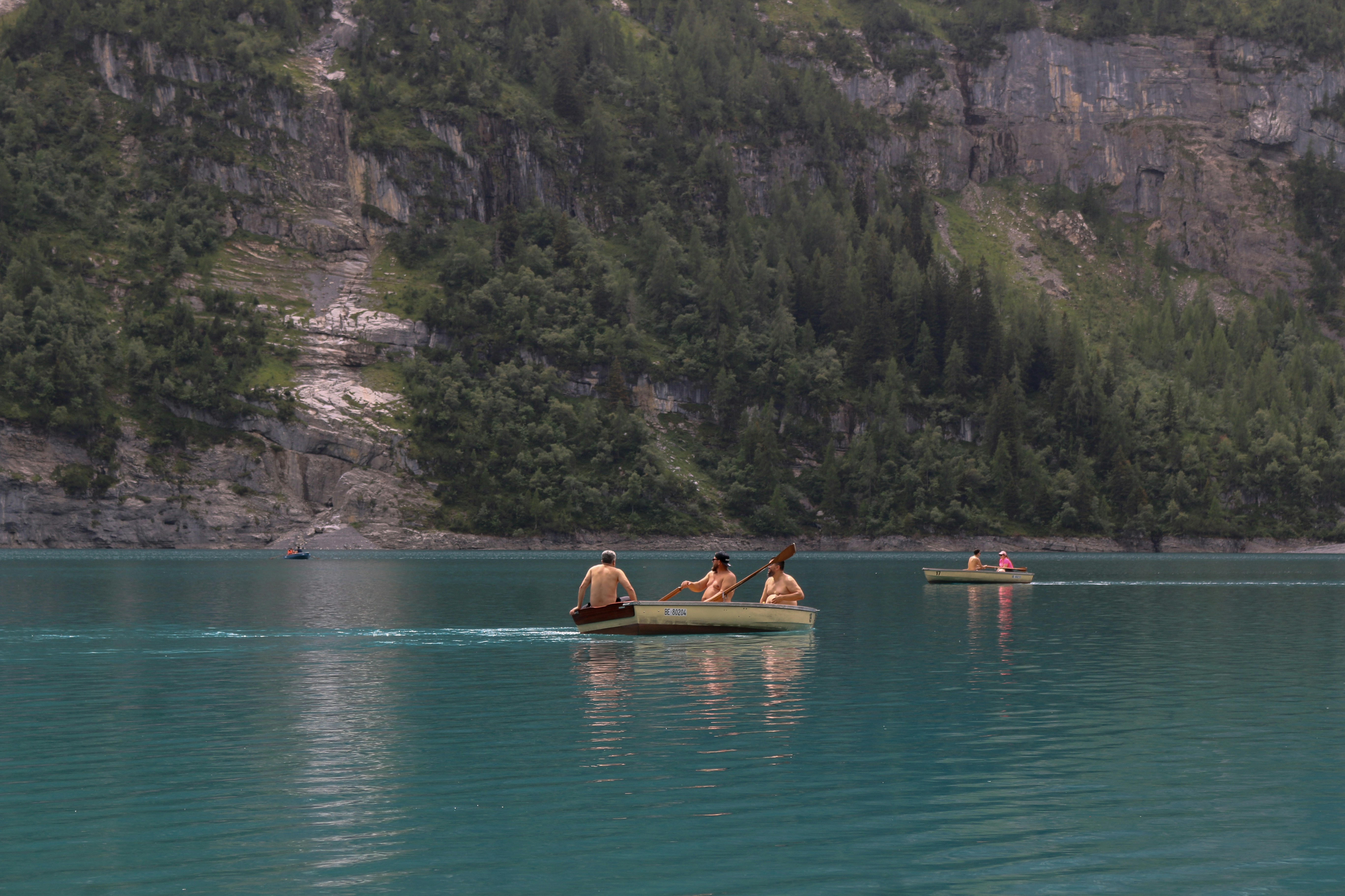 Three shirtless men rowing a small boat on a turquoise mountain lake