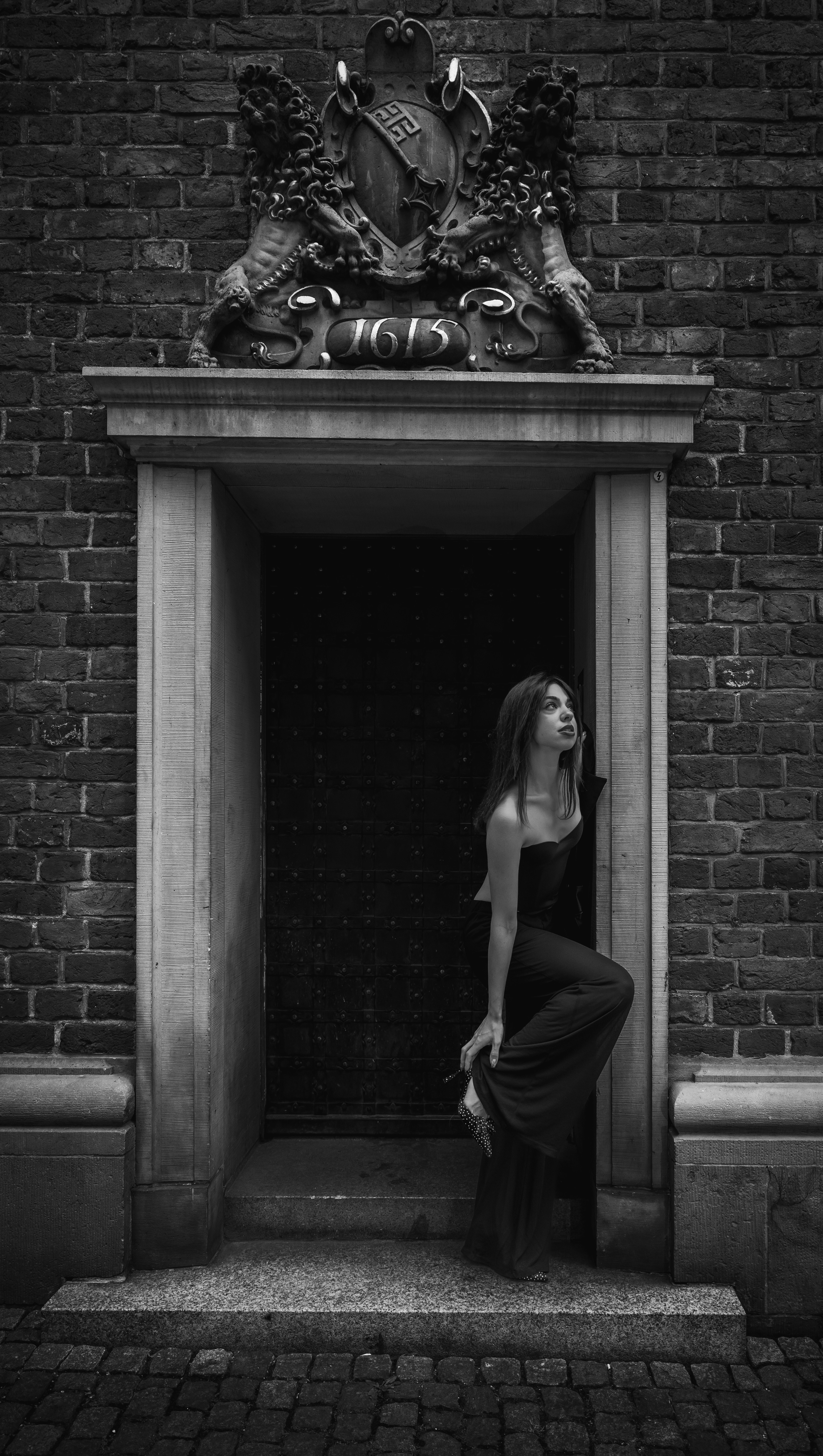 A woman in a dark jumpsuit adjusting her high heel in a stone doorway