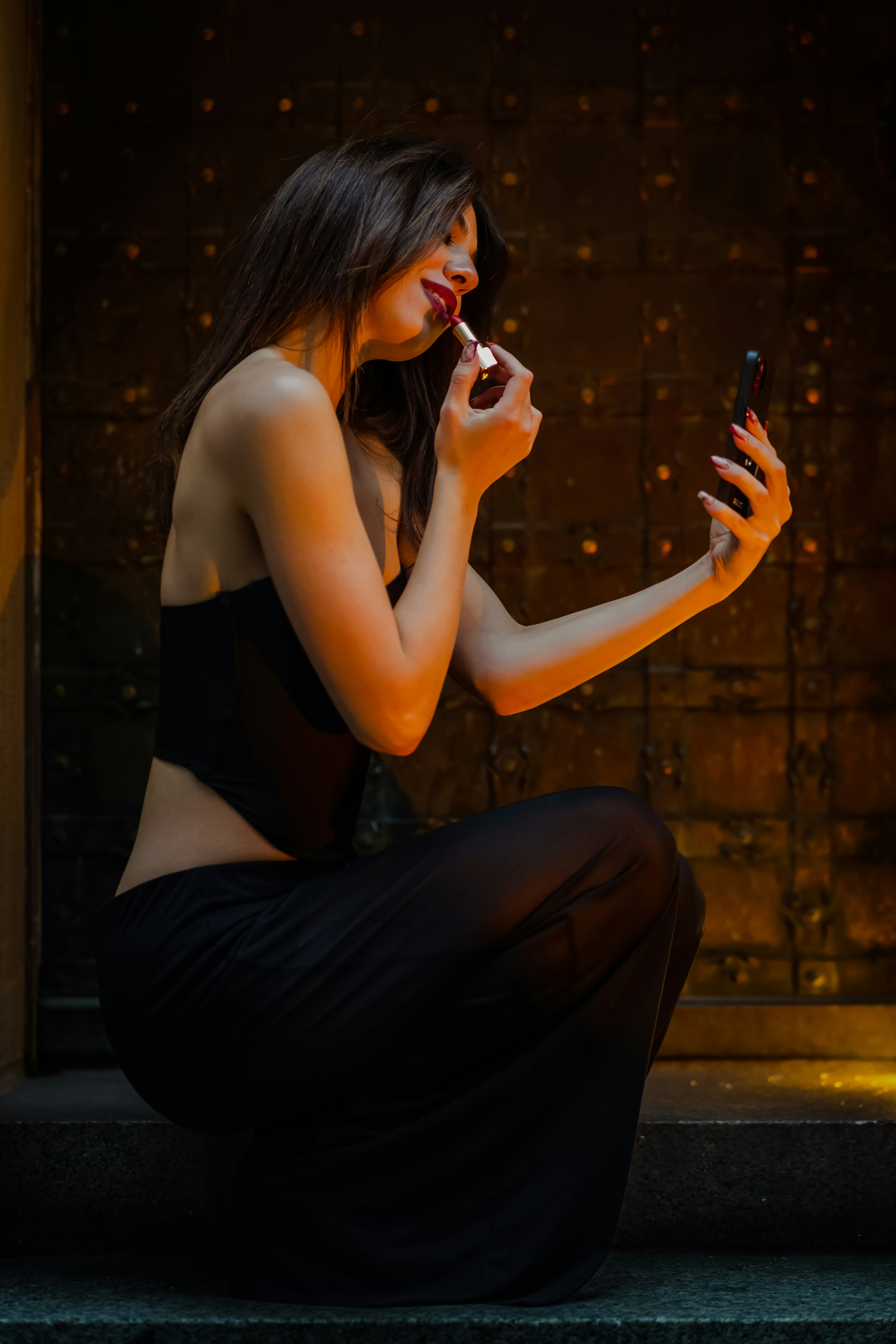A woman in a black top applying red lipstick while looking at her phone