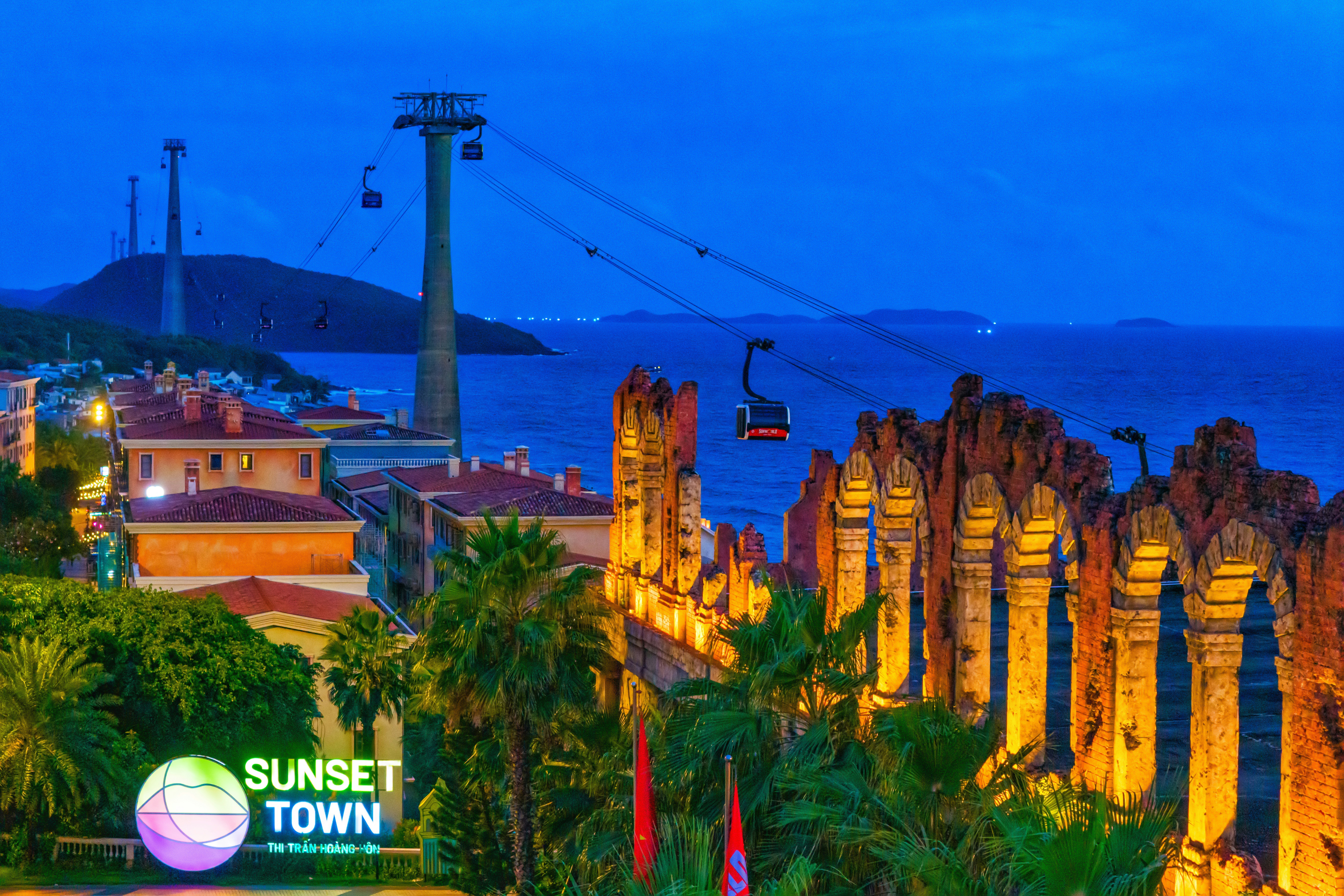 A cable car passing over stone ruins and coastal buildings at twilight in Phu Quoc