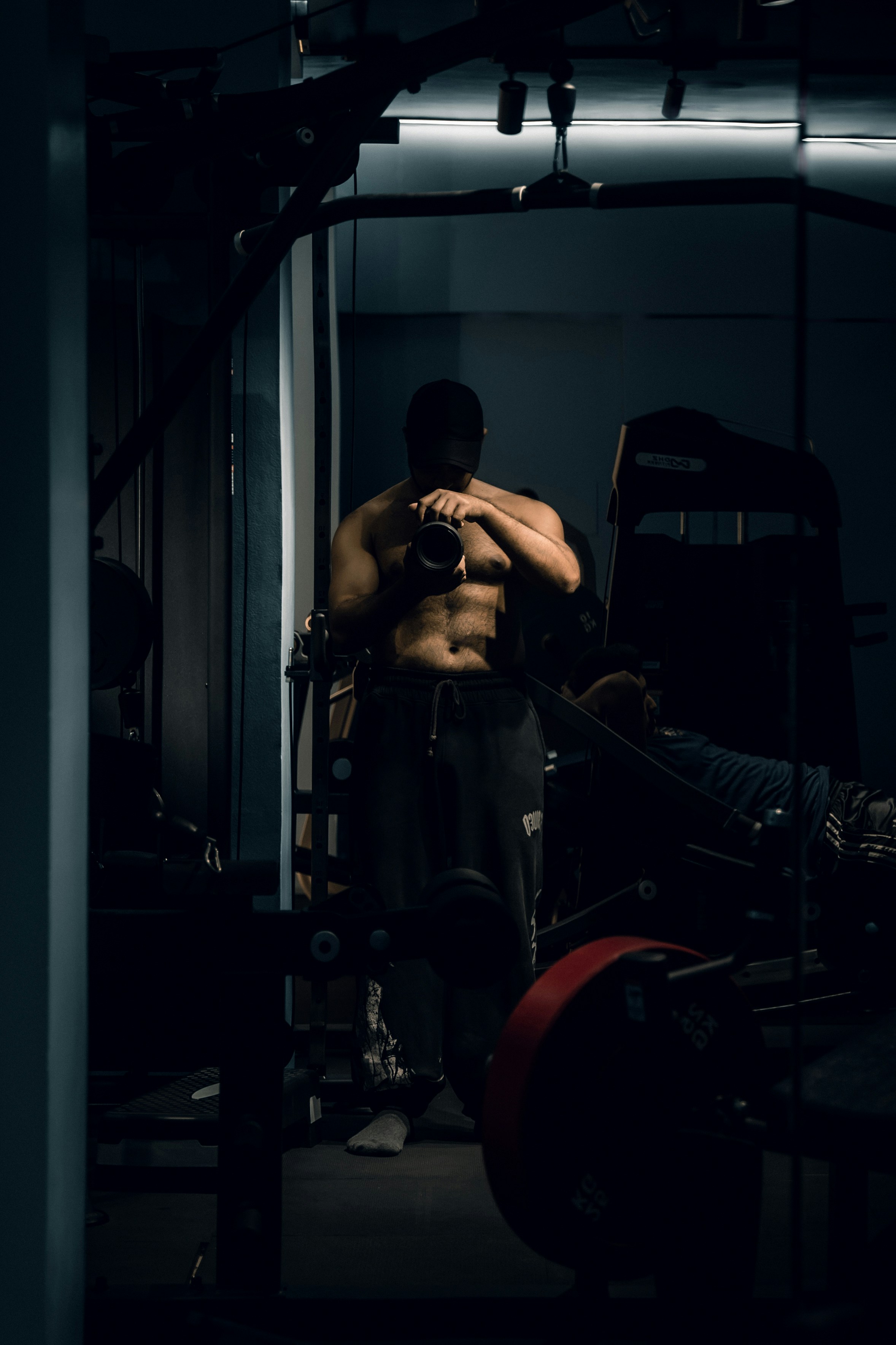 A shirtless person in a cap taking a mirror selfie in a gym