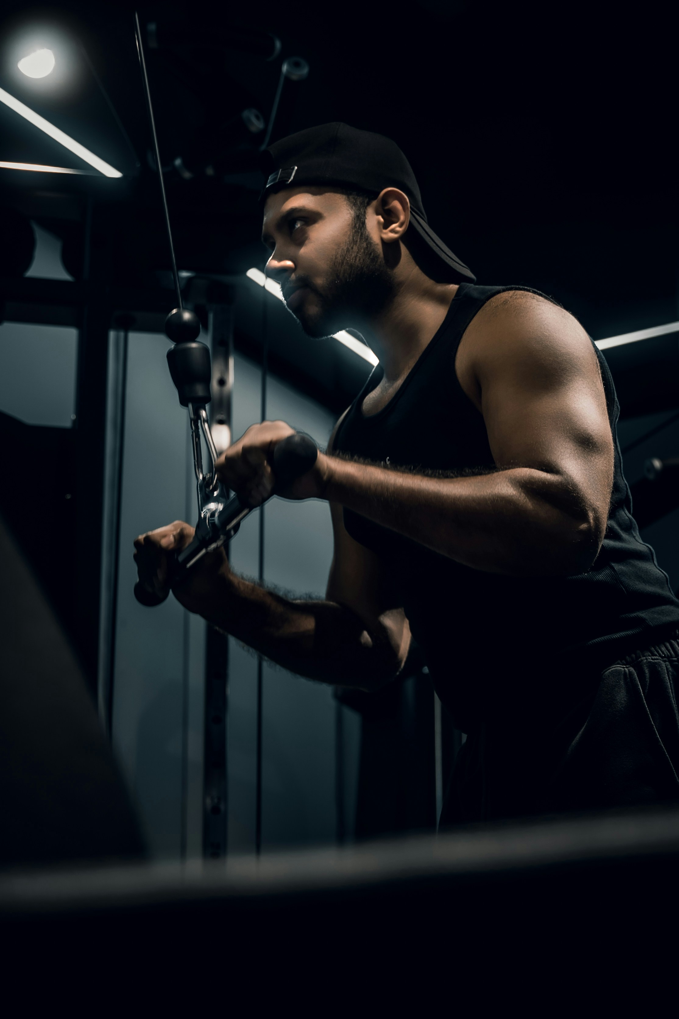 A man in a black tank top and cap performing tricep cable pushdowns