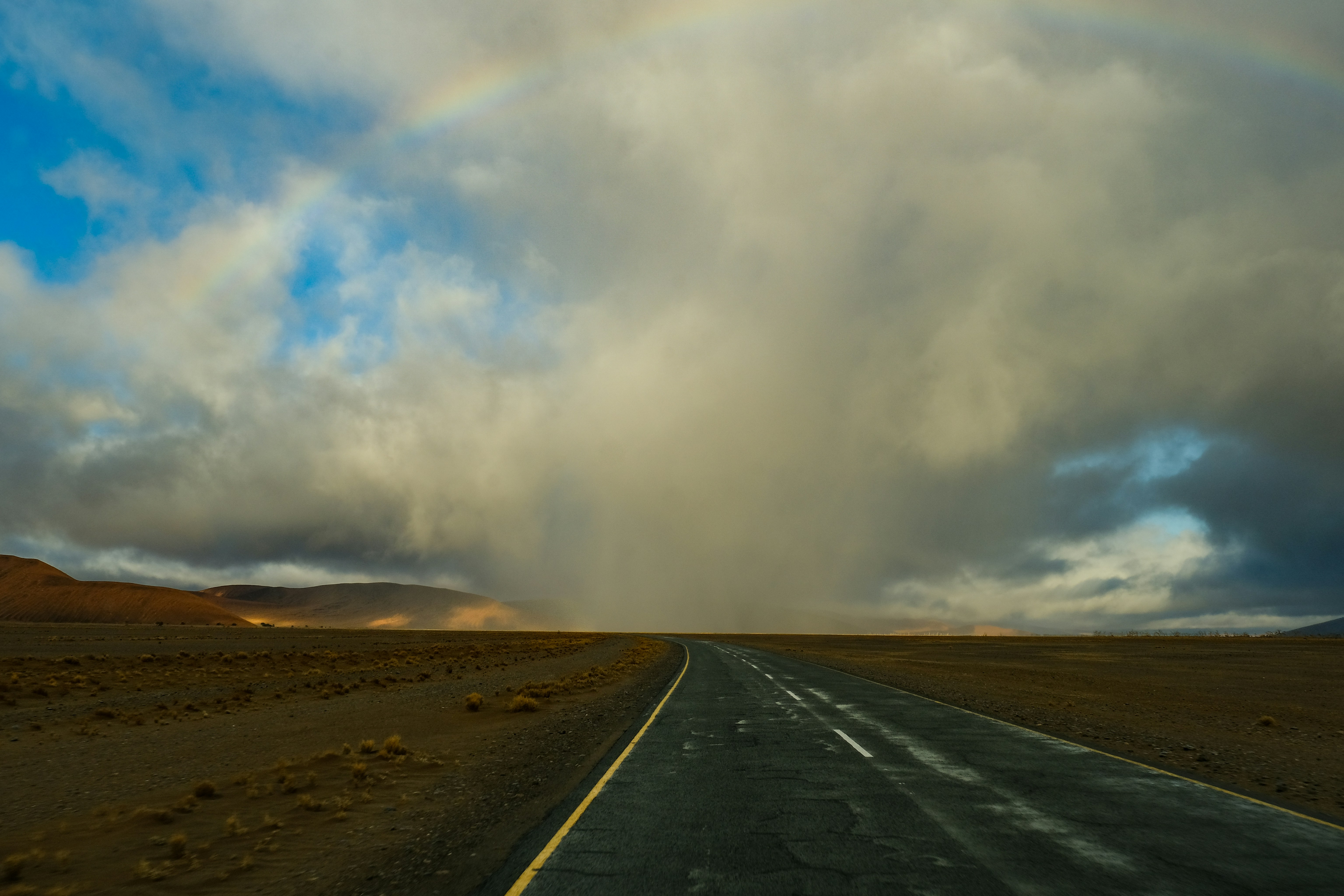 An asphalt road stretching through a vast desert landscape under a rainbow and clouds
