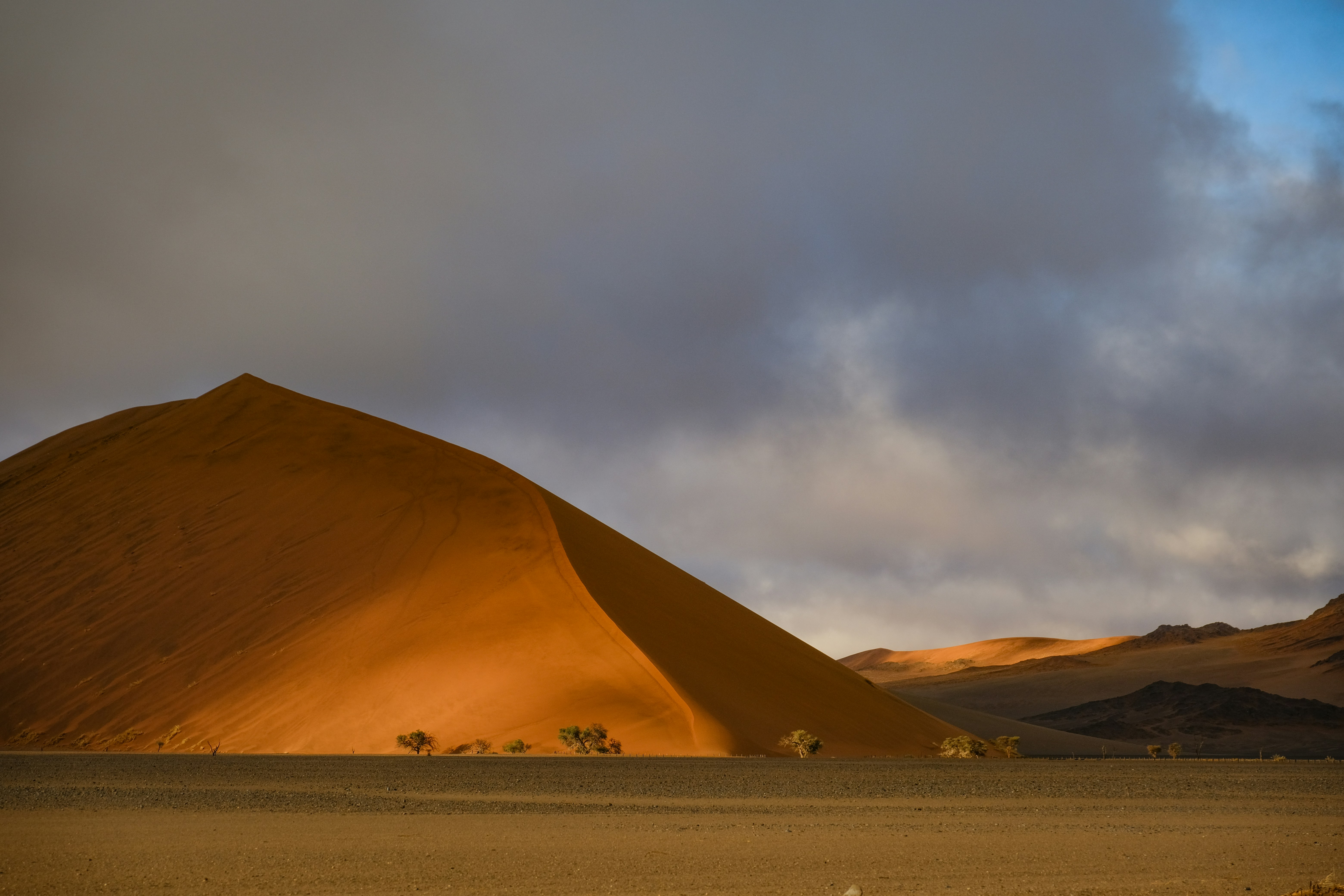 A large orange sand dune under a cloudy sky with small trees below