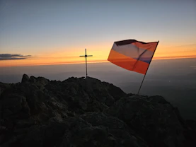 A metal cross and a tricolor flag on a rocky mountain peak at dawn