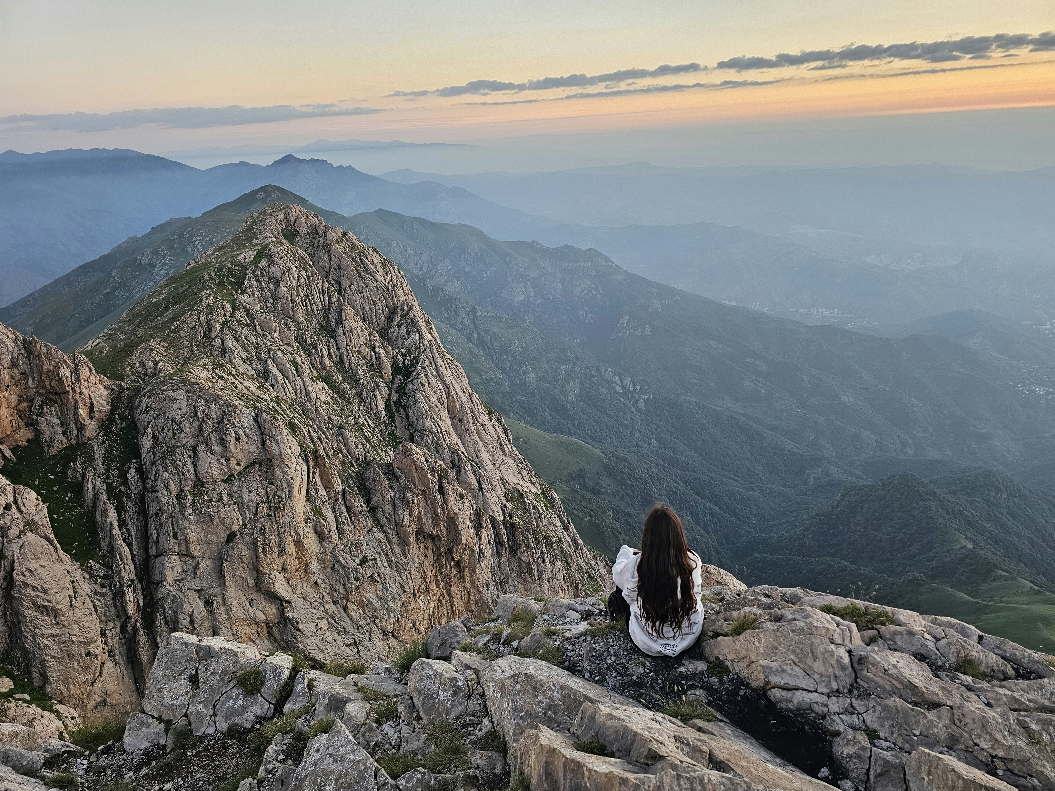 A person with long dark hair sitting on a rocky mountain peak at golden hour