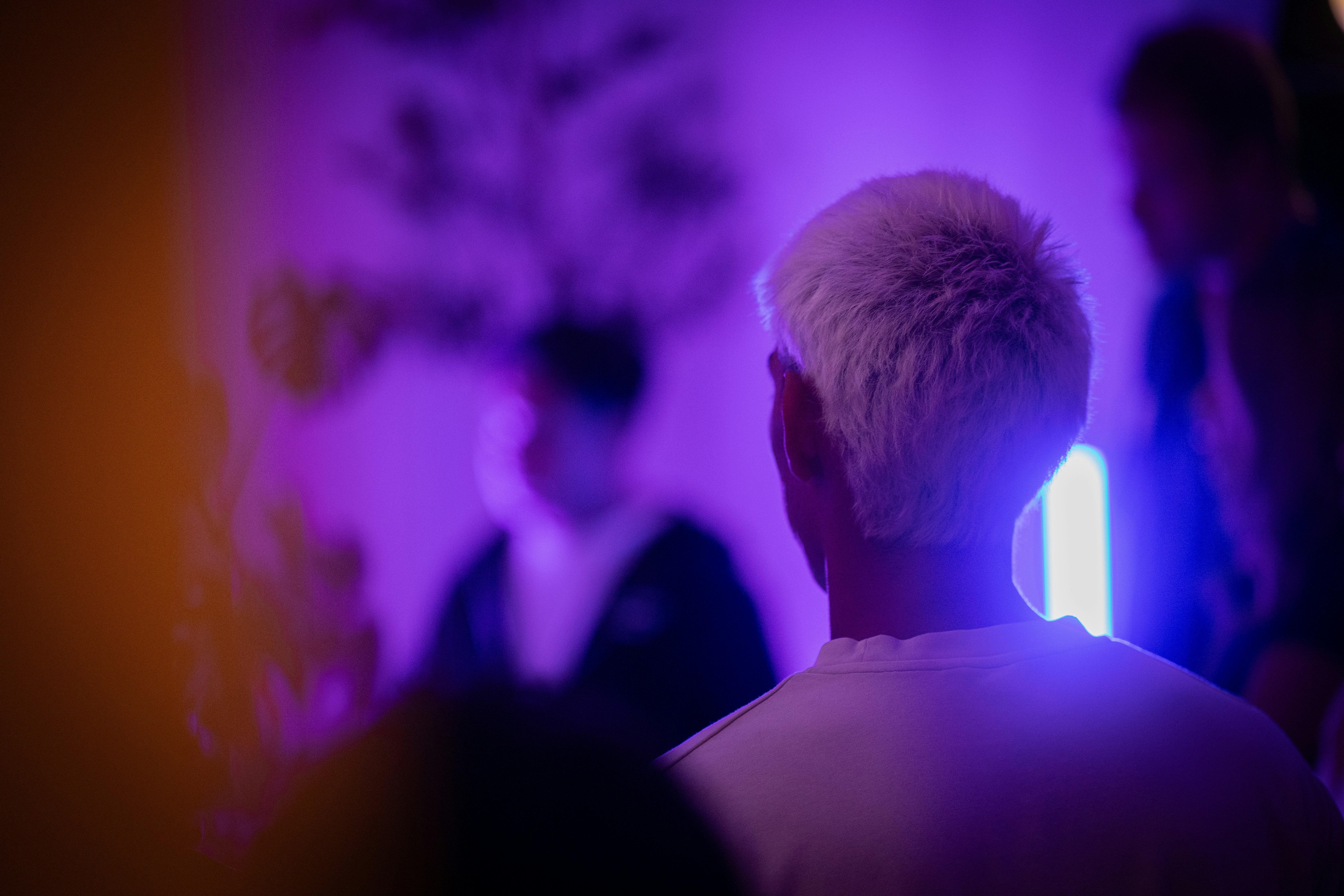A person with short bleached hair in a white shirt under purple light