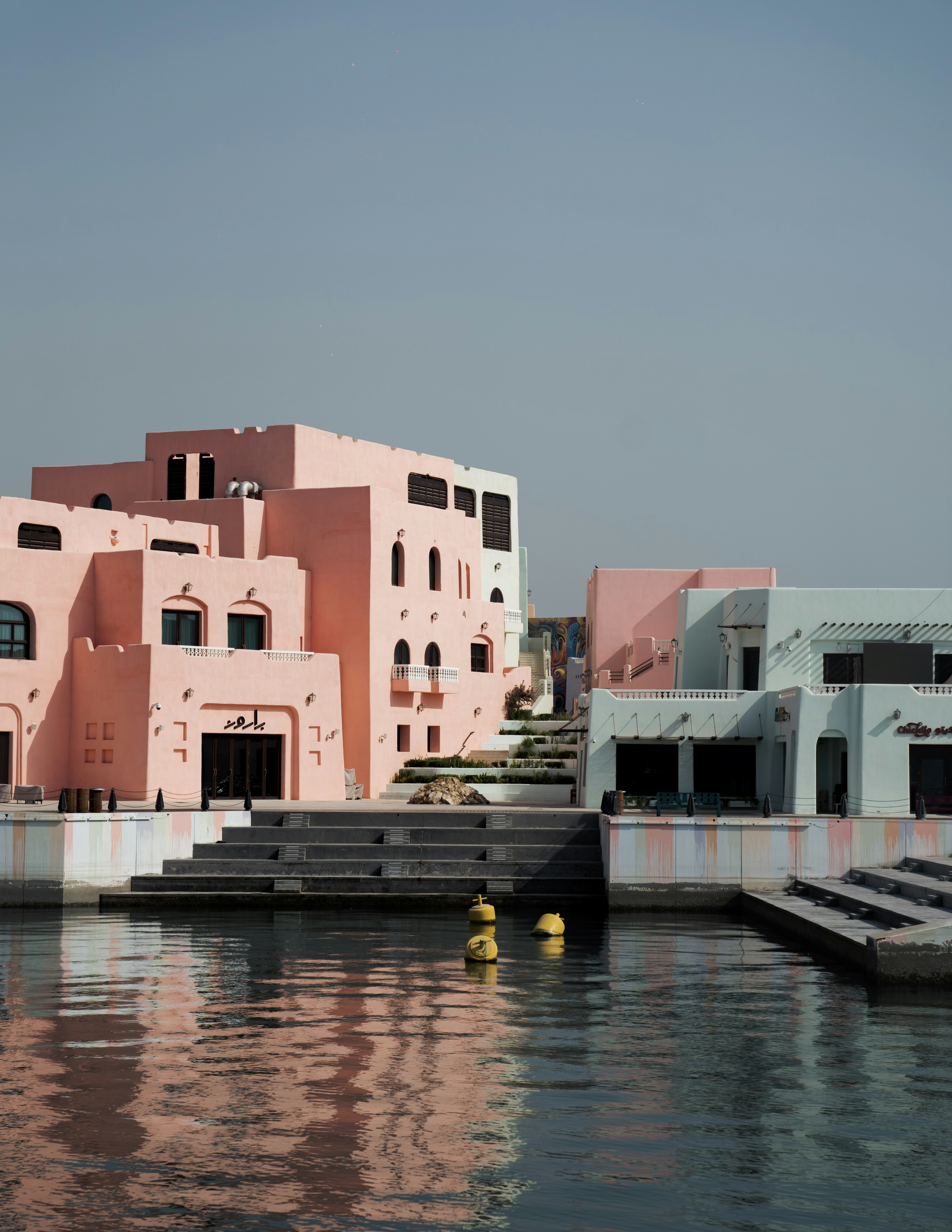 Pink and light blue buildings along a waterfront with stone steps and yellow buoys