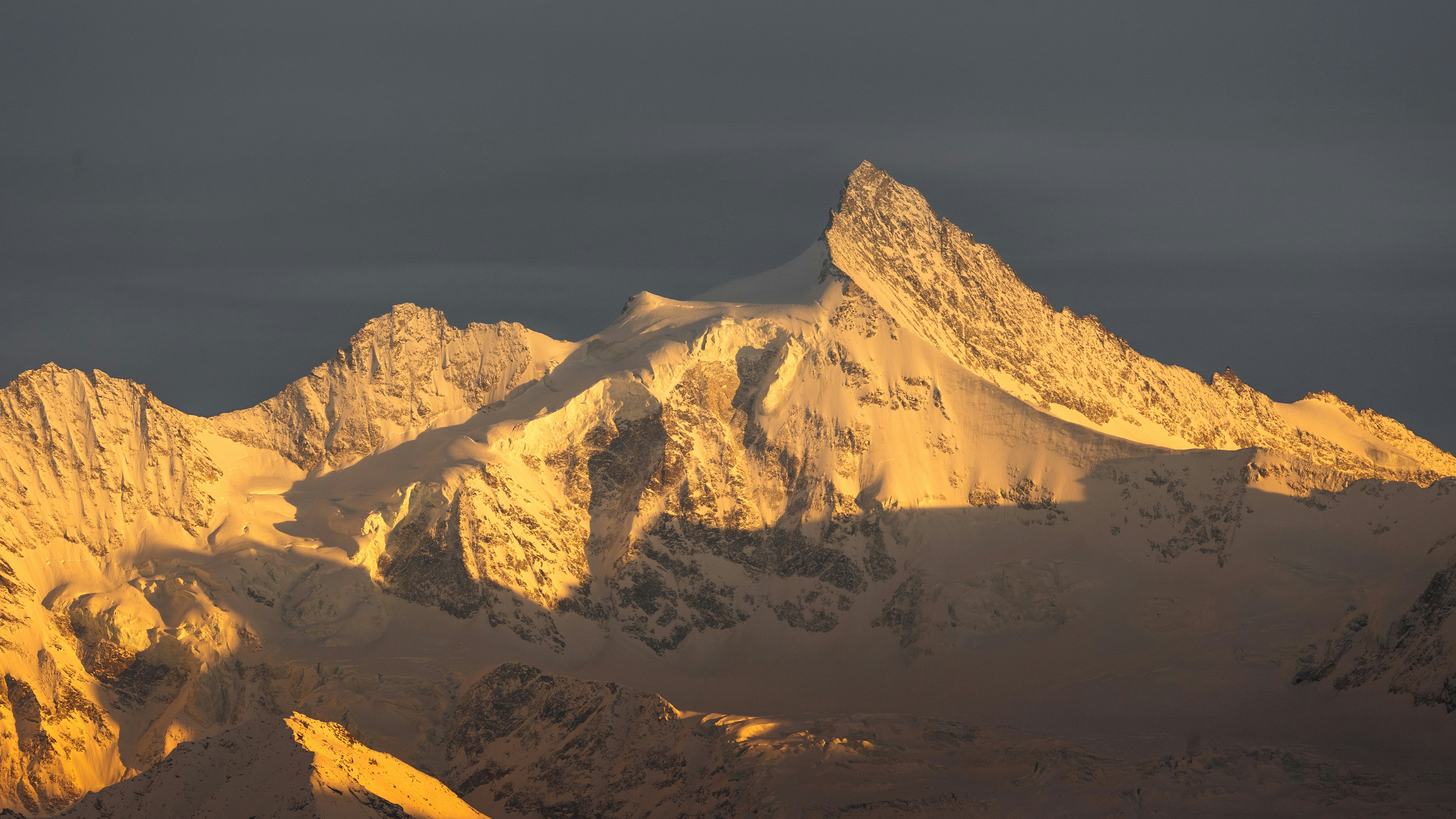 Snowy mountain peaks illuminated by warm golden light against a dark grey sky