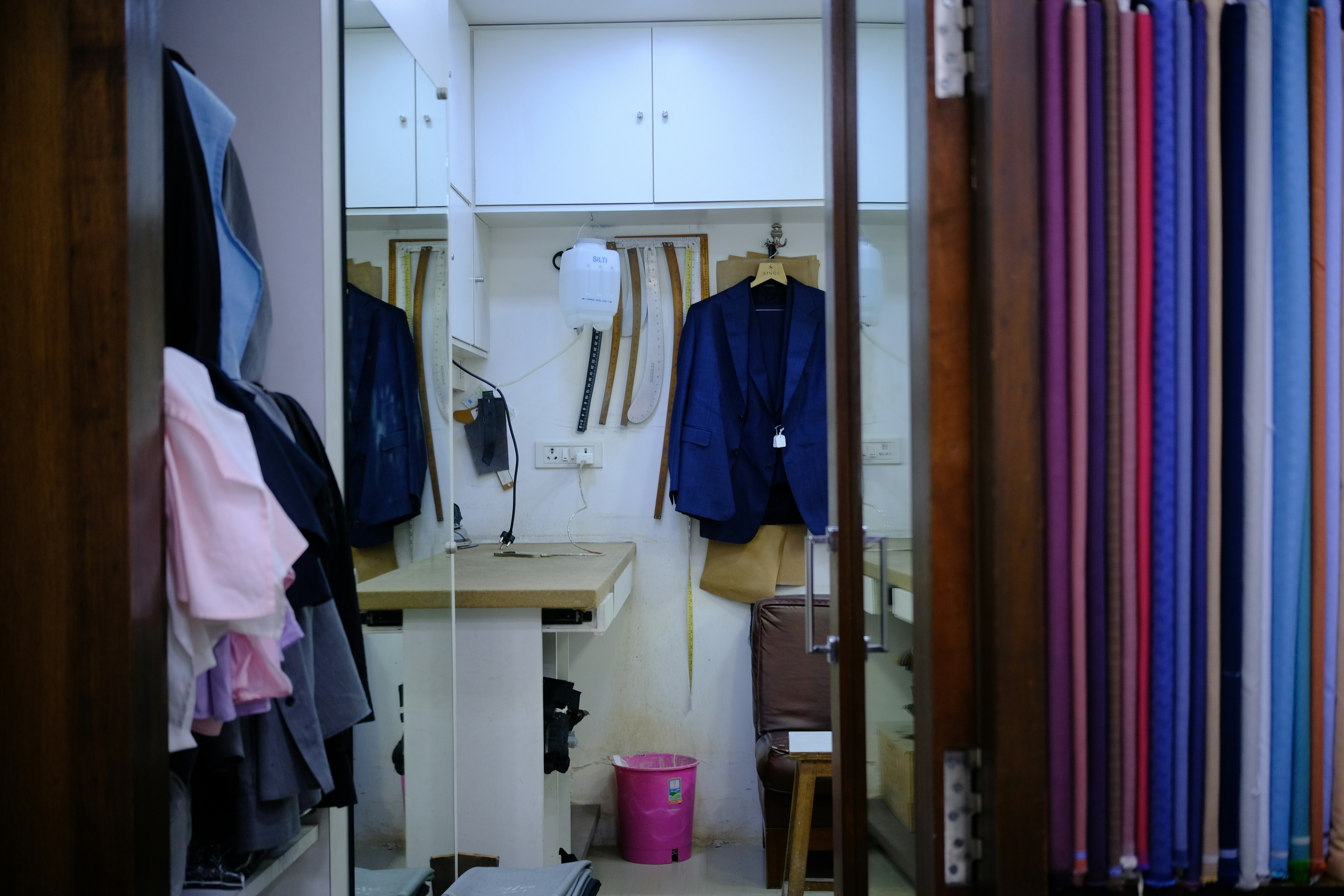 A tailor shop interior with hanging suits, fabric rolls, and a workbench