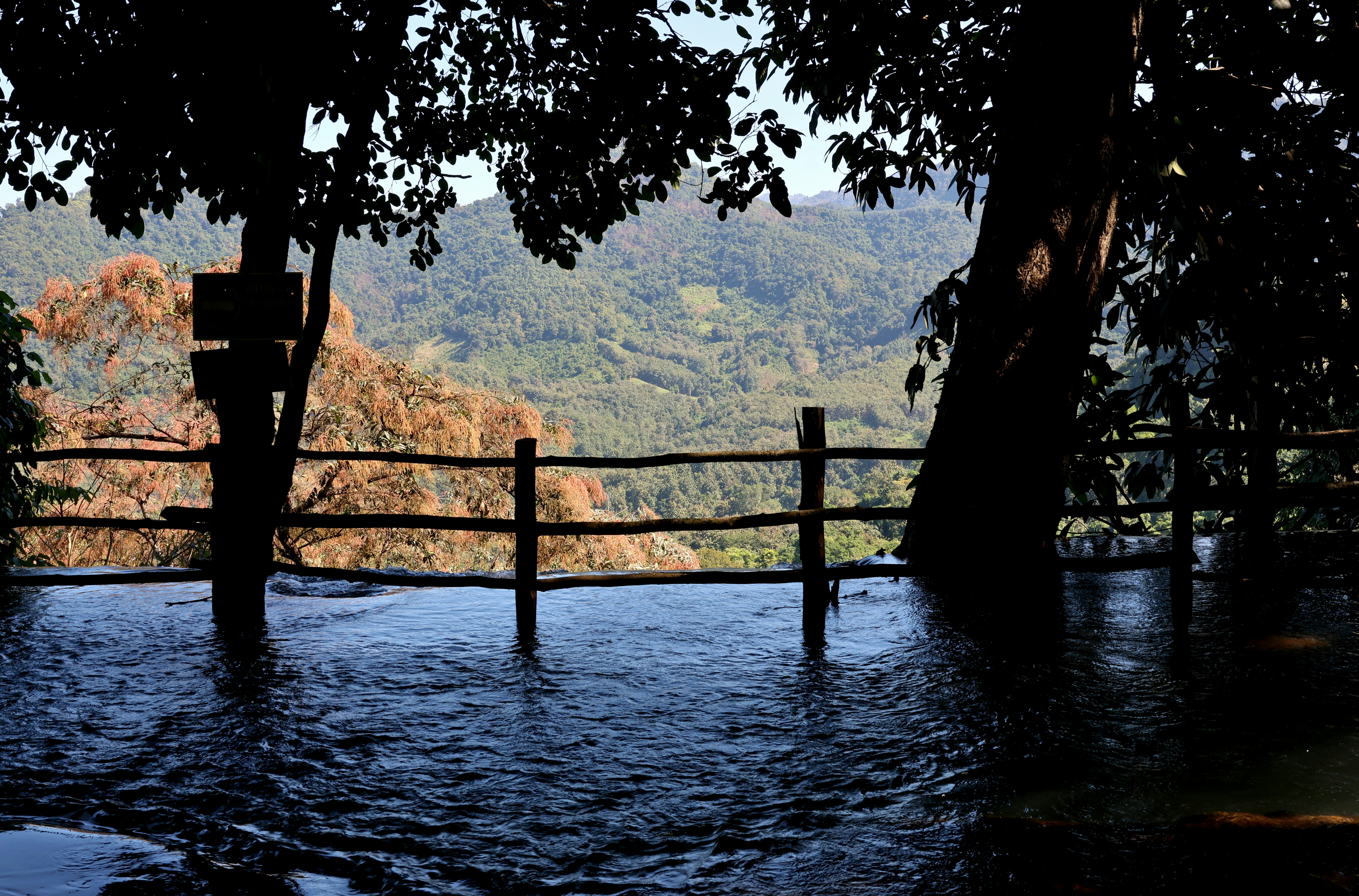 Laos, December 2025, a wooden fence at the edge of a flowing stream overlooking forested mountains