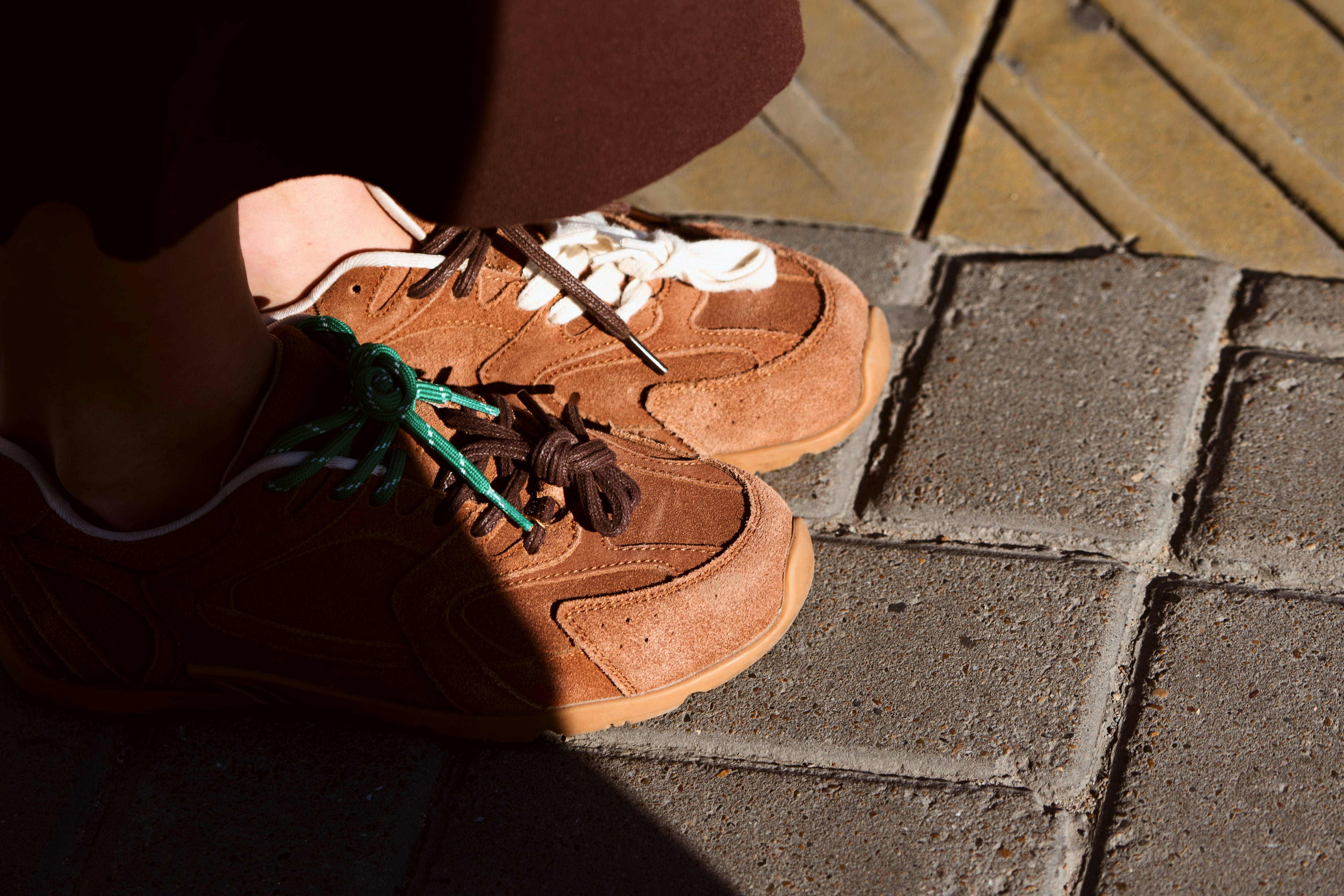 Brown suede sneakers with mismatched green and brown laces on a stone sidewalk