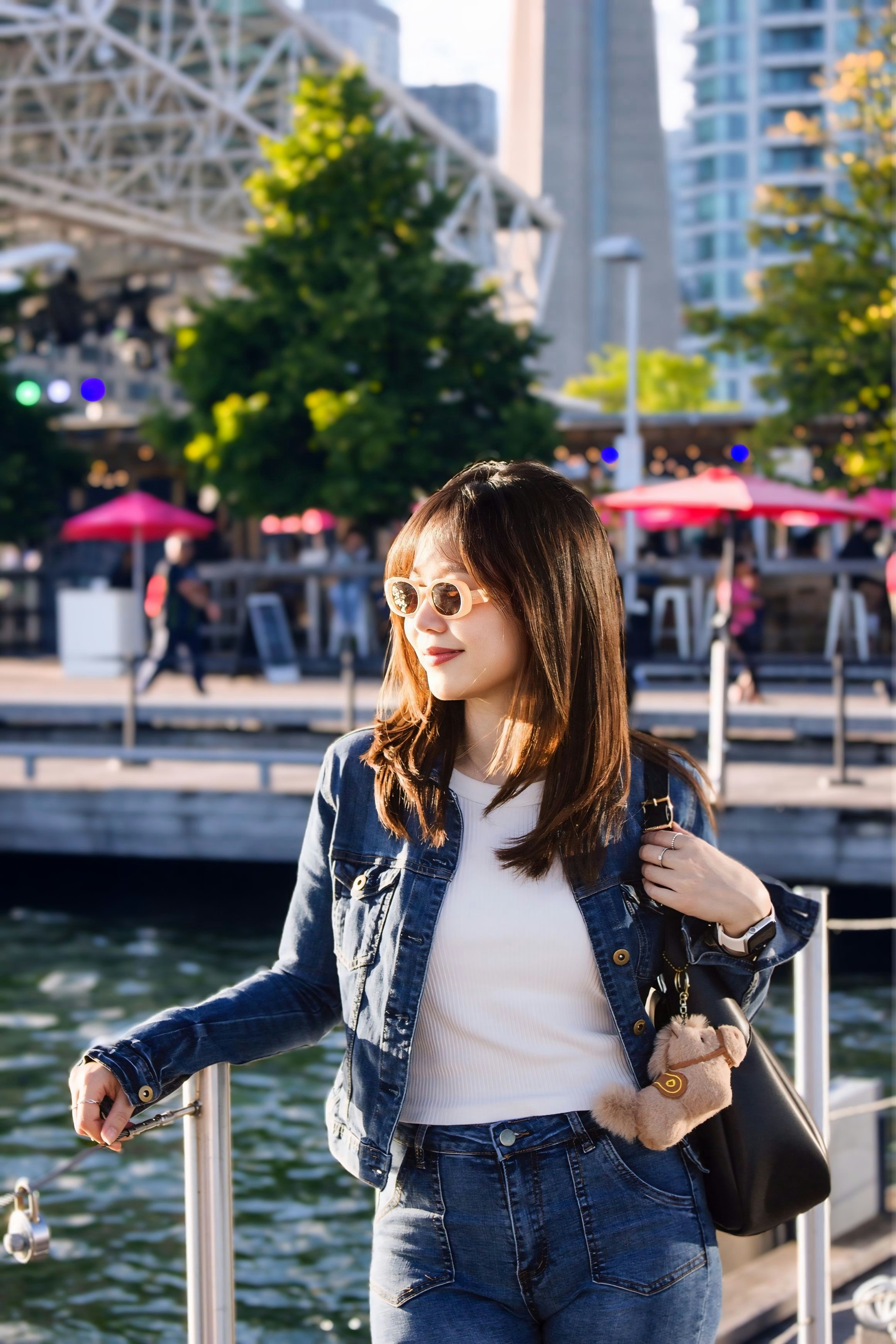 A woman in a denim jacket and sunglasses standing by a waterfront promenade