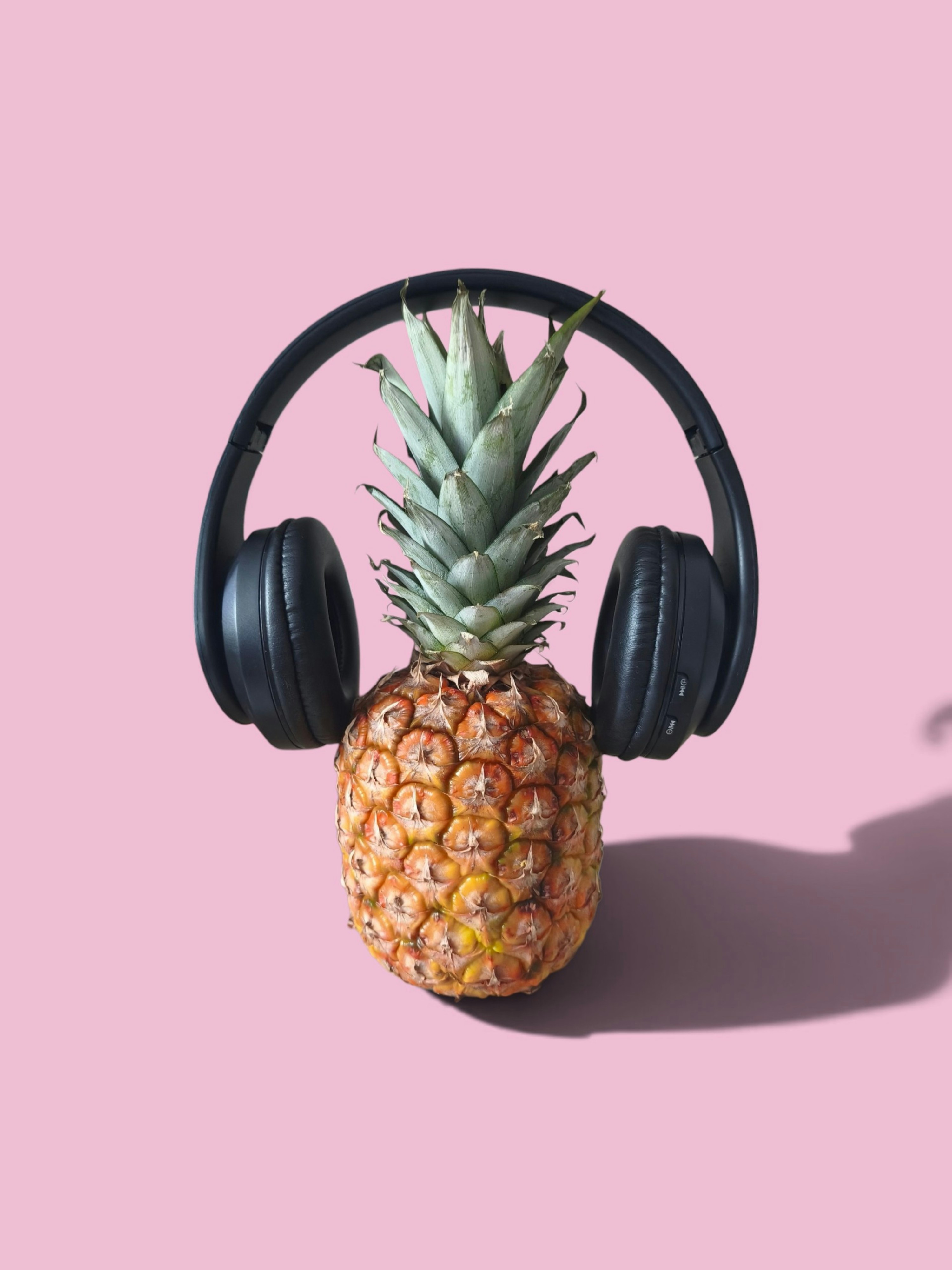 A whole pineapple wearing black over-ear headphones against a solid pink background