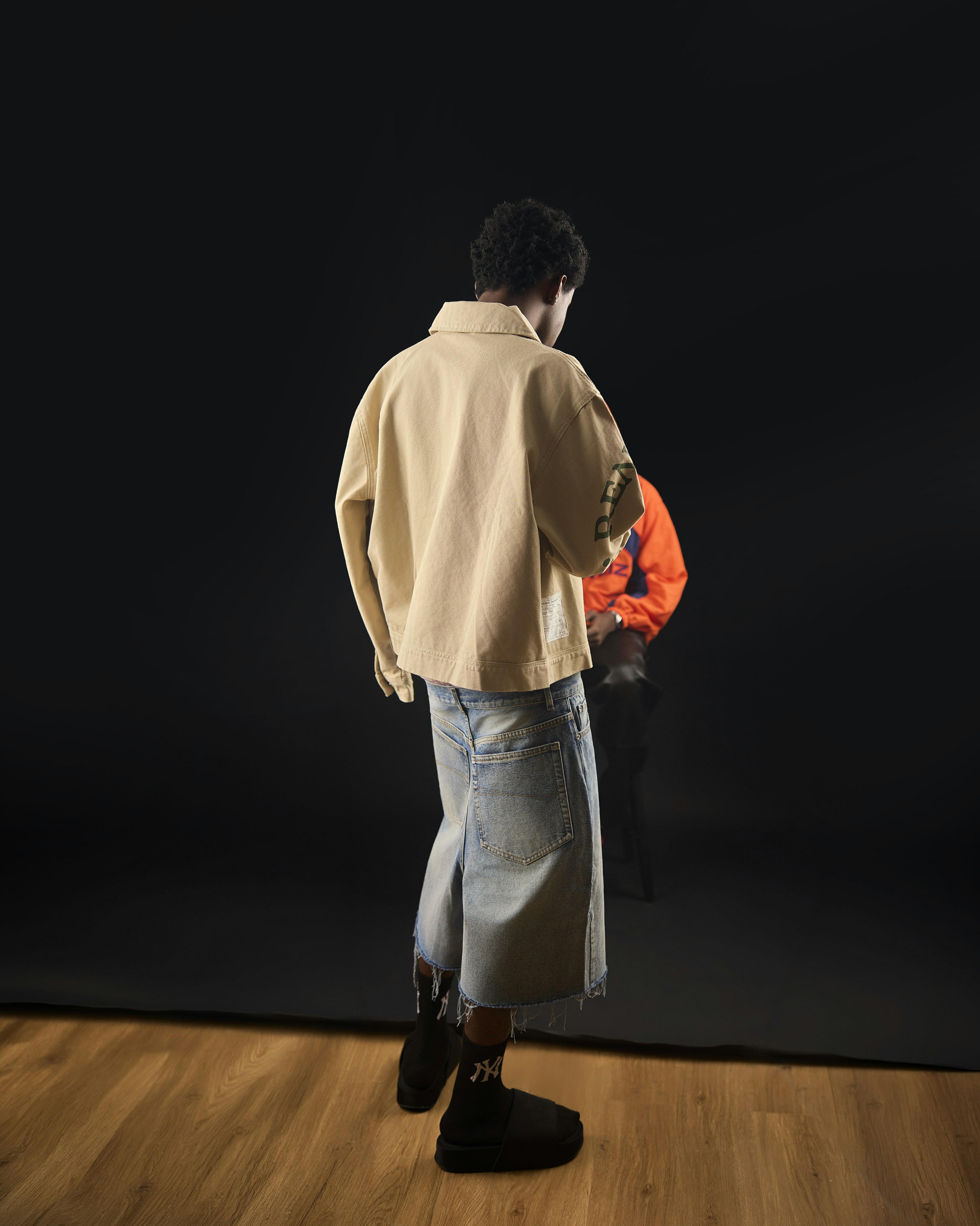 A person in a beige jacket and denim shorts facing a seated person