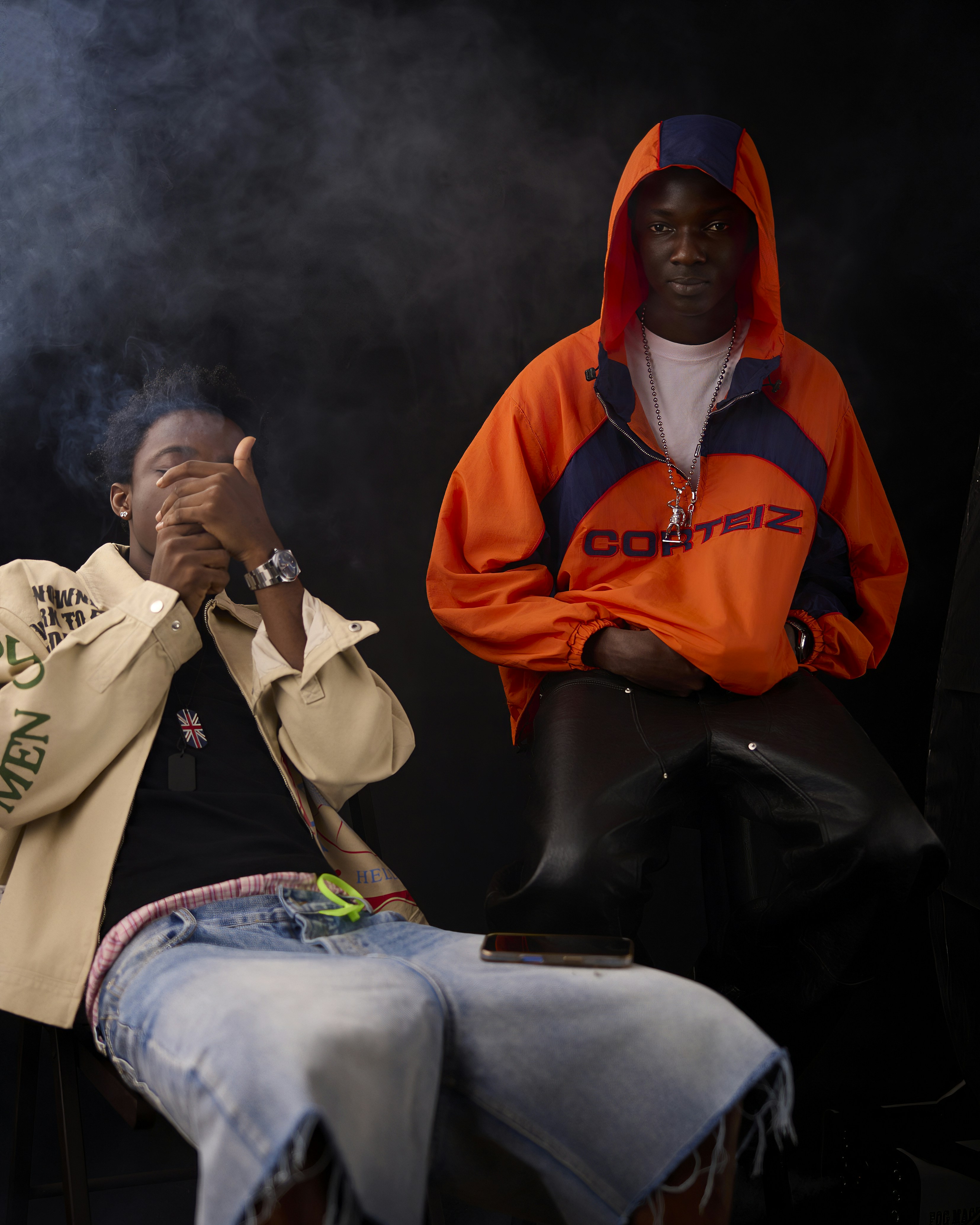 Two men in streetwear against a black background with smoke, one lighting a cigarette