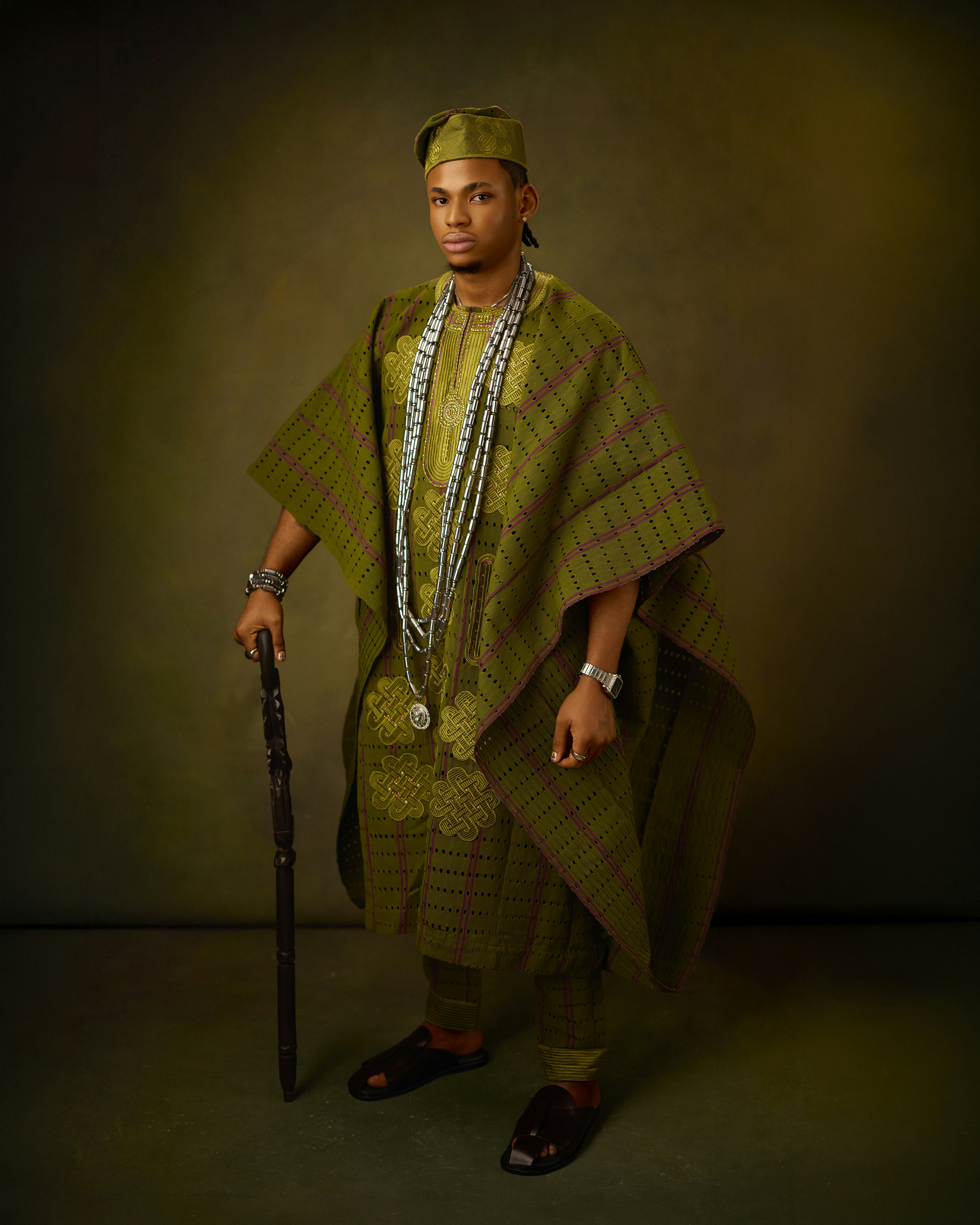 A man in a green patterned agbada and cap holding a wooden walking stick