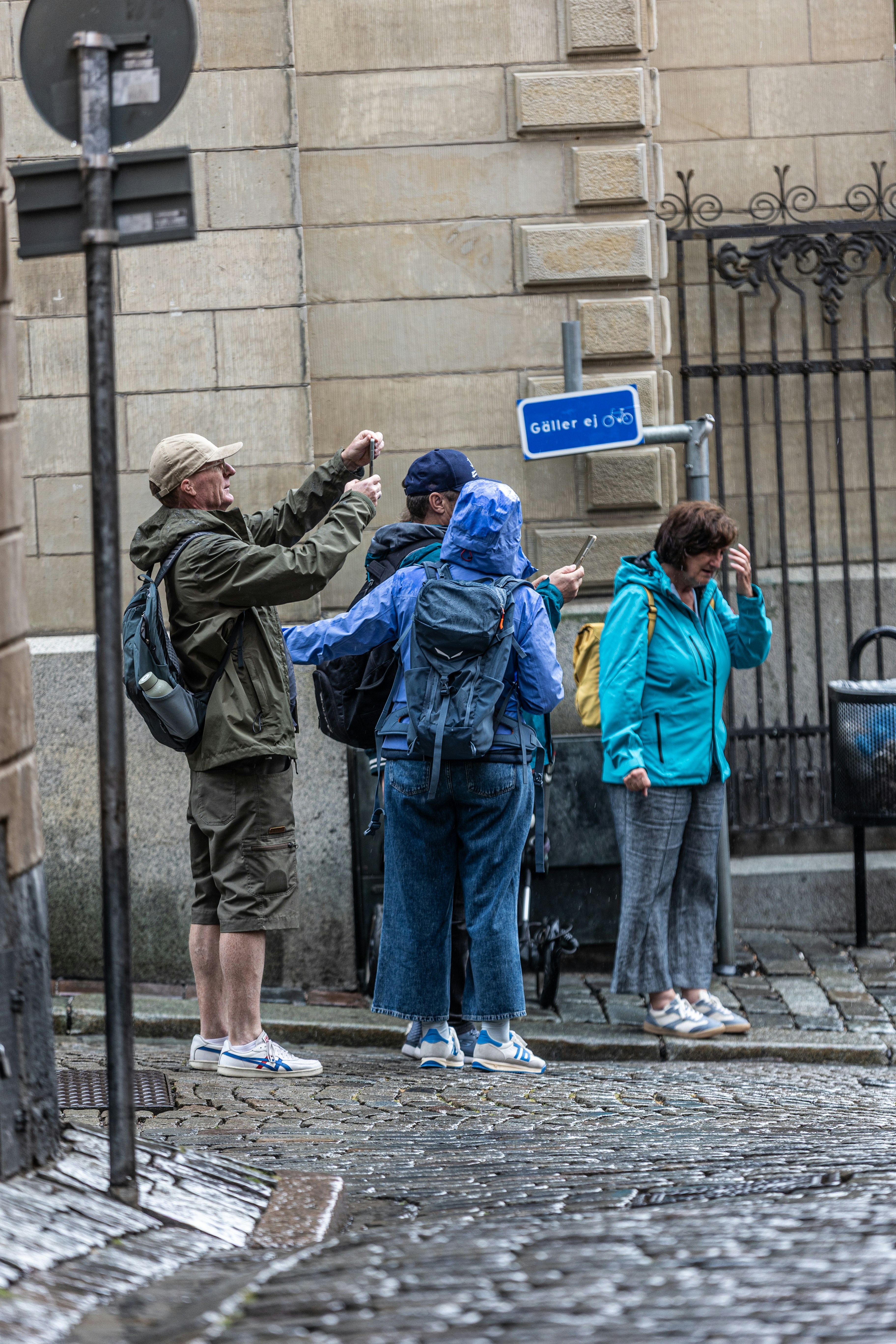 Three people in rain gear taking photos on a wet cobblestone street