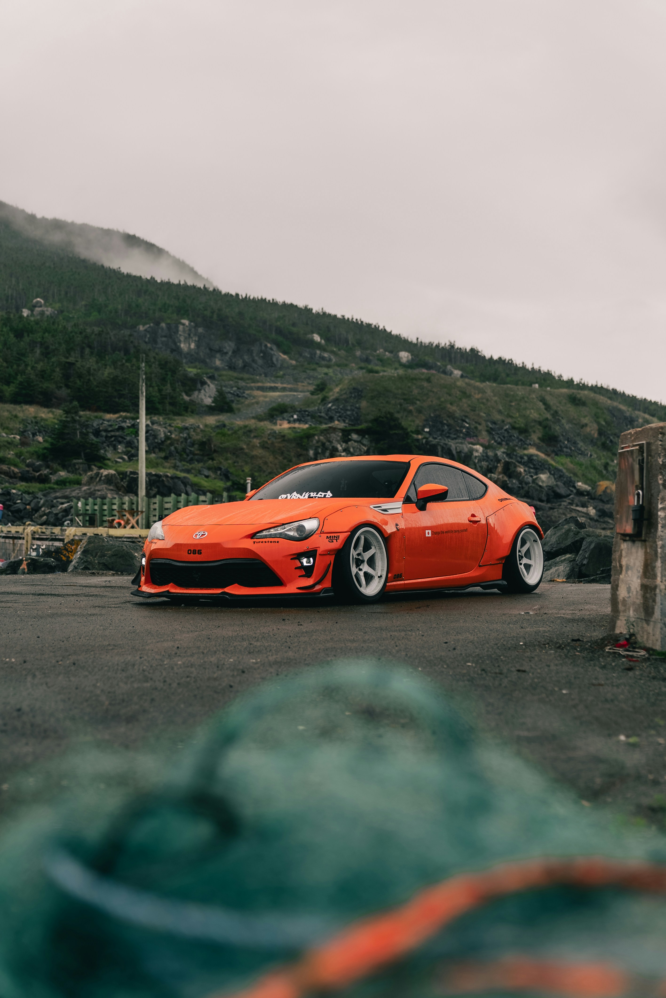 An orange sports car with wide fenders parked on a wet coastal road