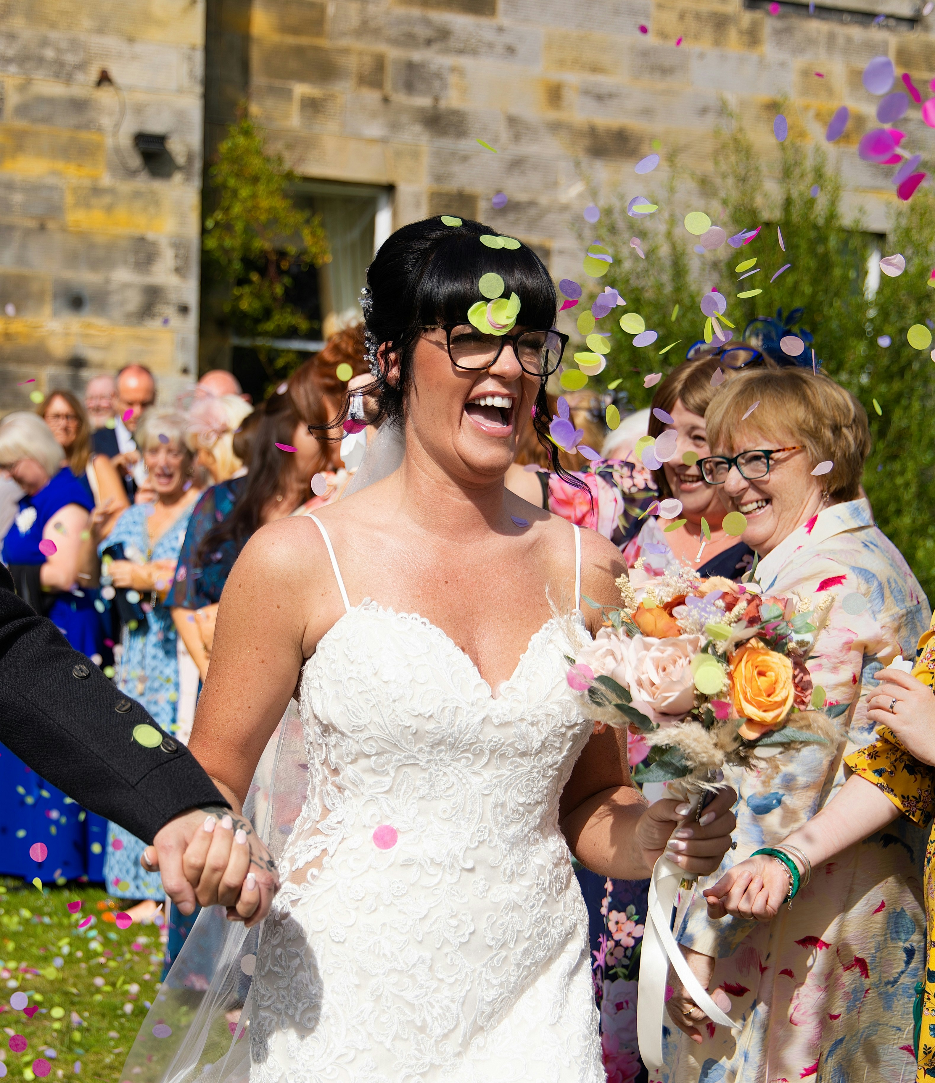 A laughing bride in a white lace dress holding a bouquet amidst falling confetti