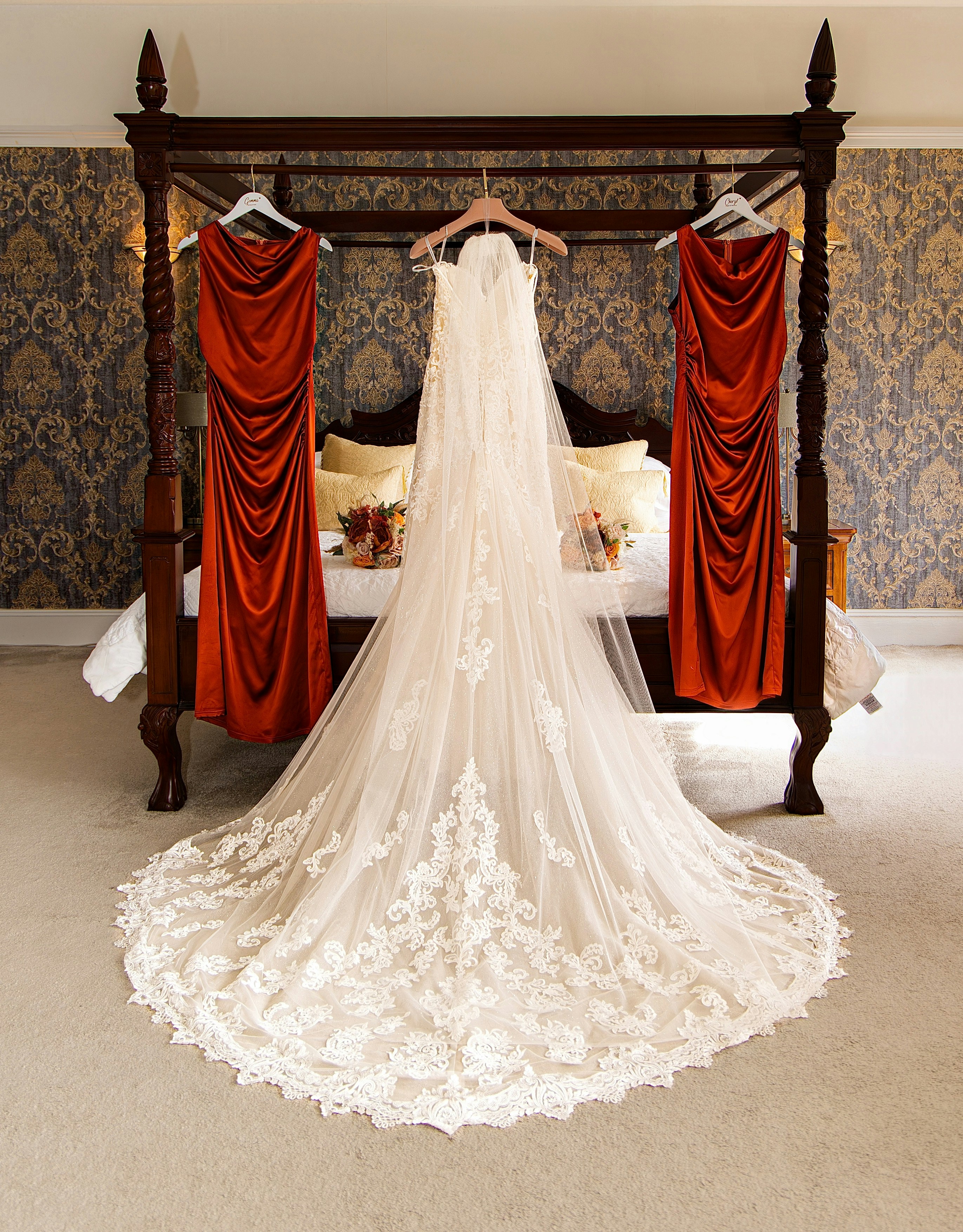 A white wedding dress and two rust-colored bridesmaid dresses hanging on a four-poster bed