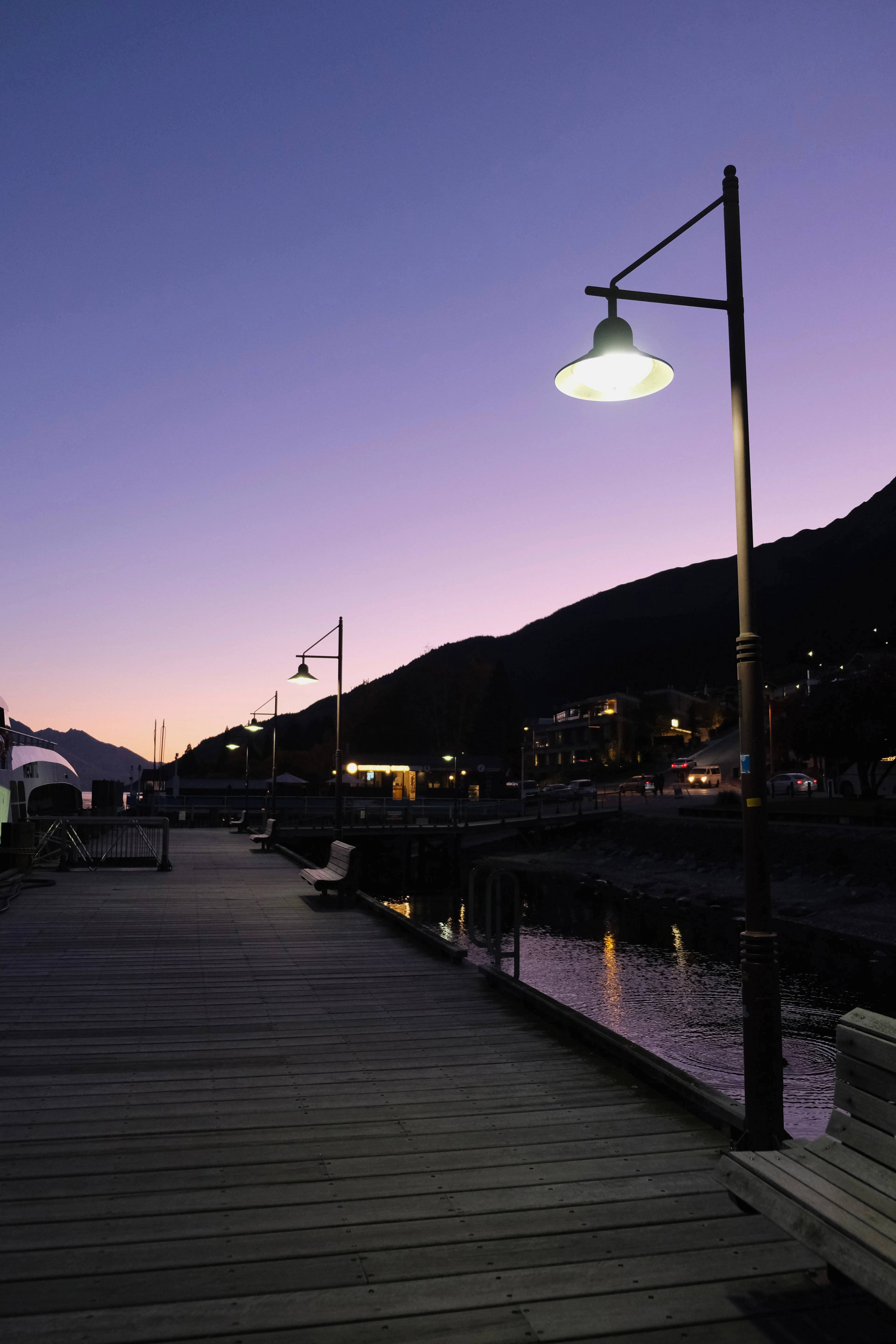 The wooden waterfront boardwalk in Queenstown after sunset, its lamps burning under a violet and rose sky, with the lights of the town reflected in the still water of Lake Wakatipu.