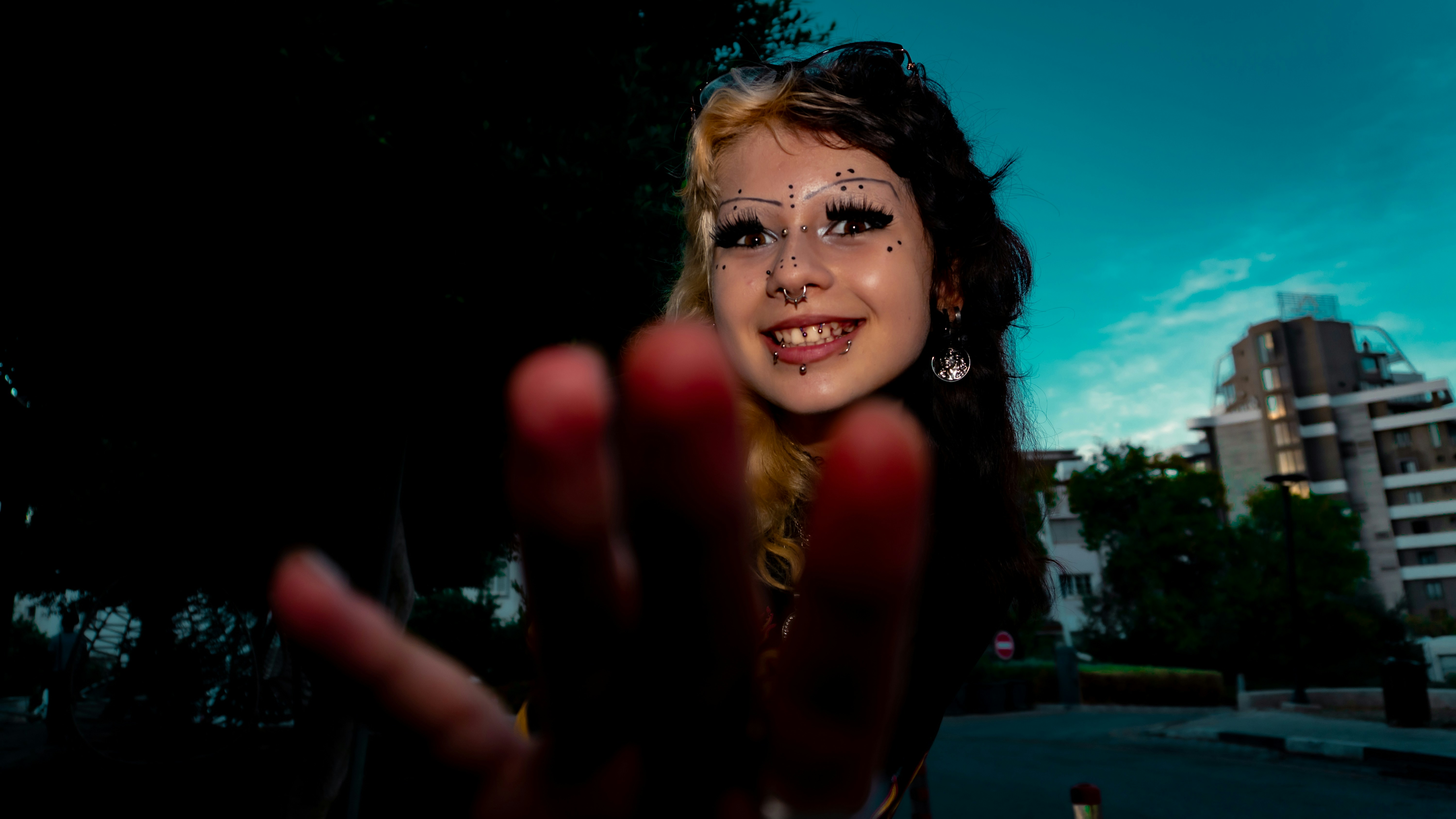 A person with two-toned hair and facial piercings reaching toward the camera outdoors