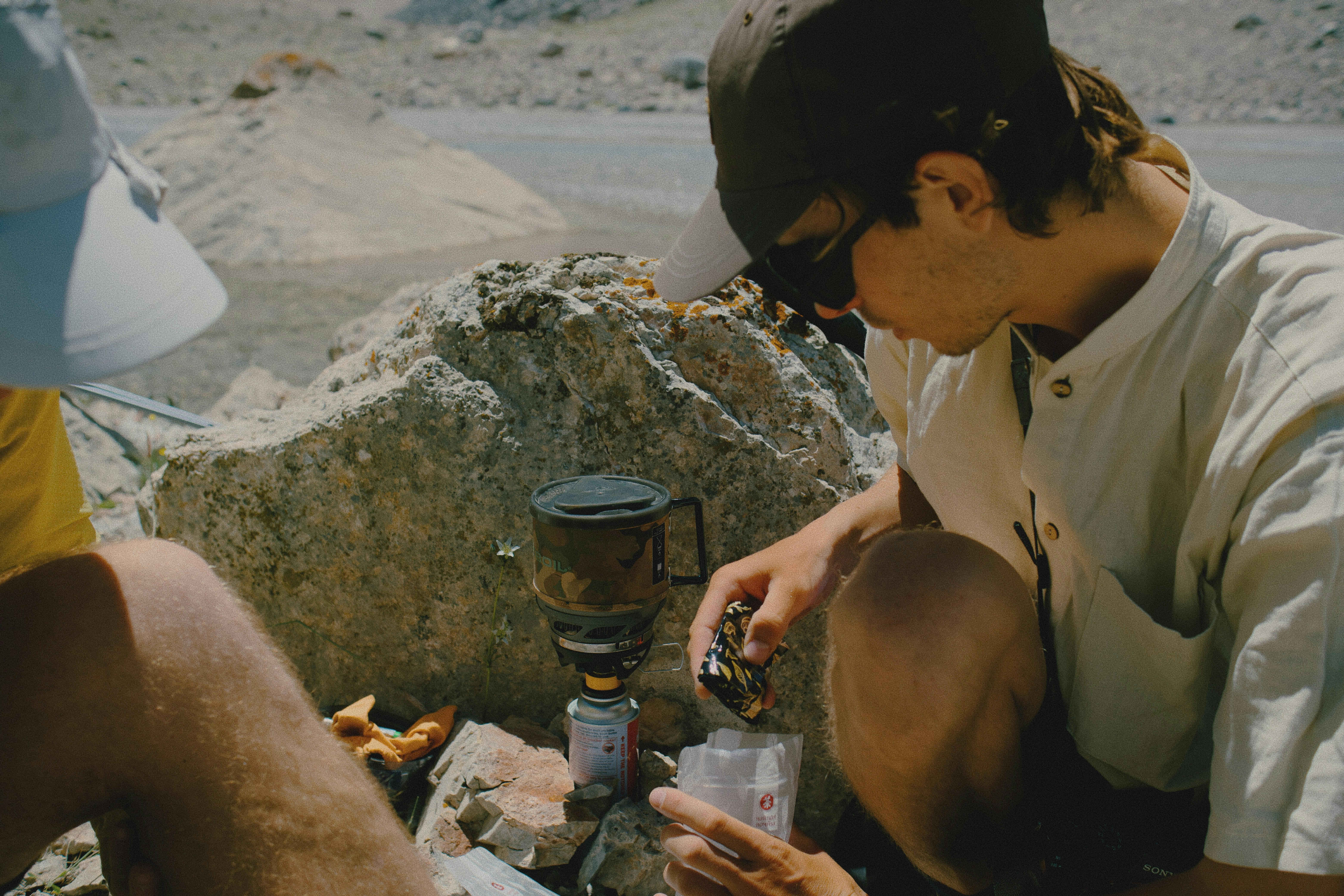 A person in a cap and sunglasses preparing food on a camping stove