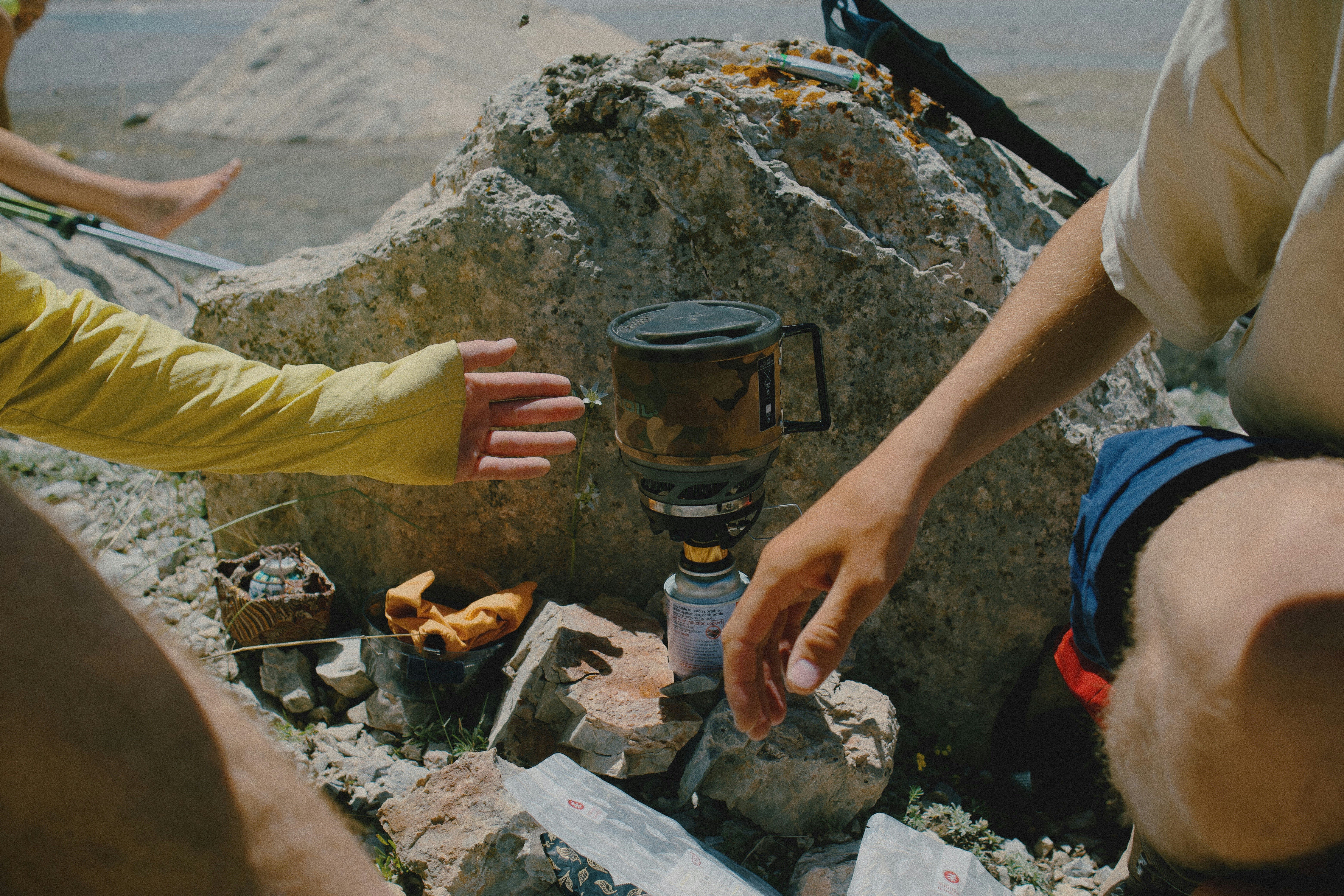 People reaching toward a portable camping stove on a rocky surface outdoors