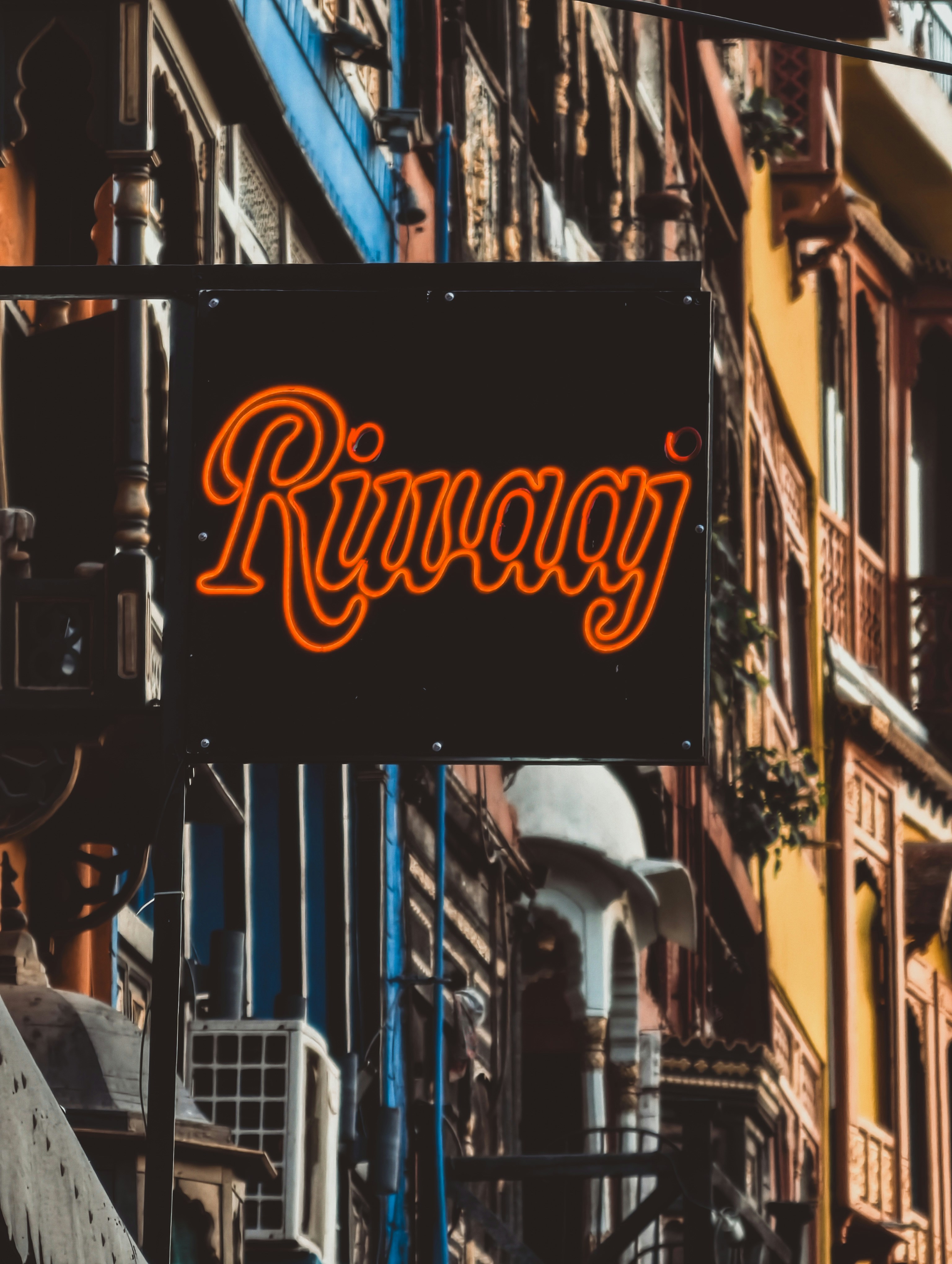 A black sign with orange neon lettering reading Riwaaj on a city street