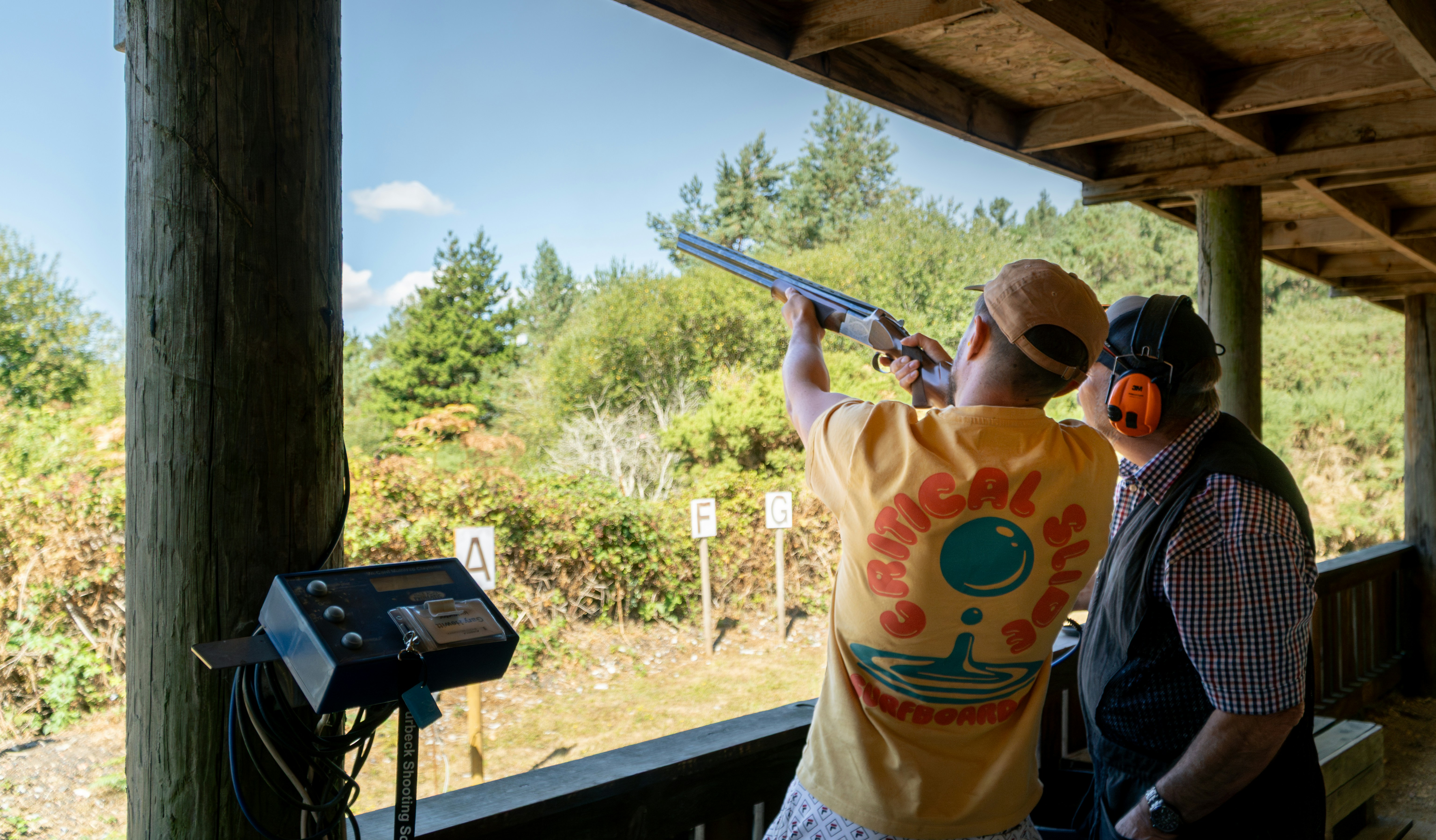 A person in a yellow t-shirt aiming a shotgun at an outdoor shooting range