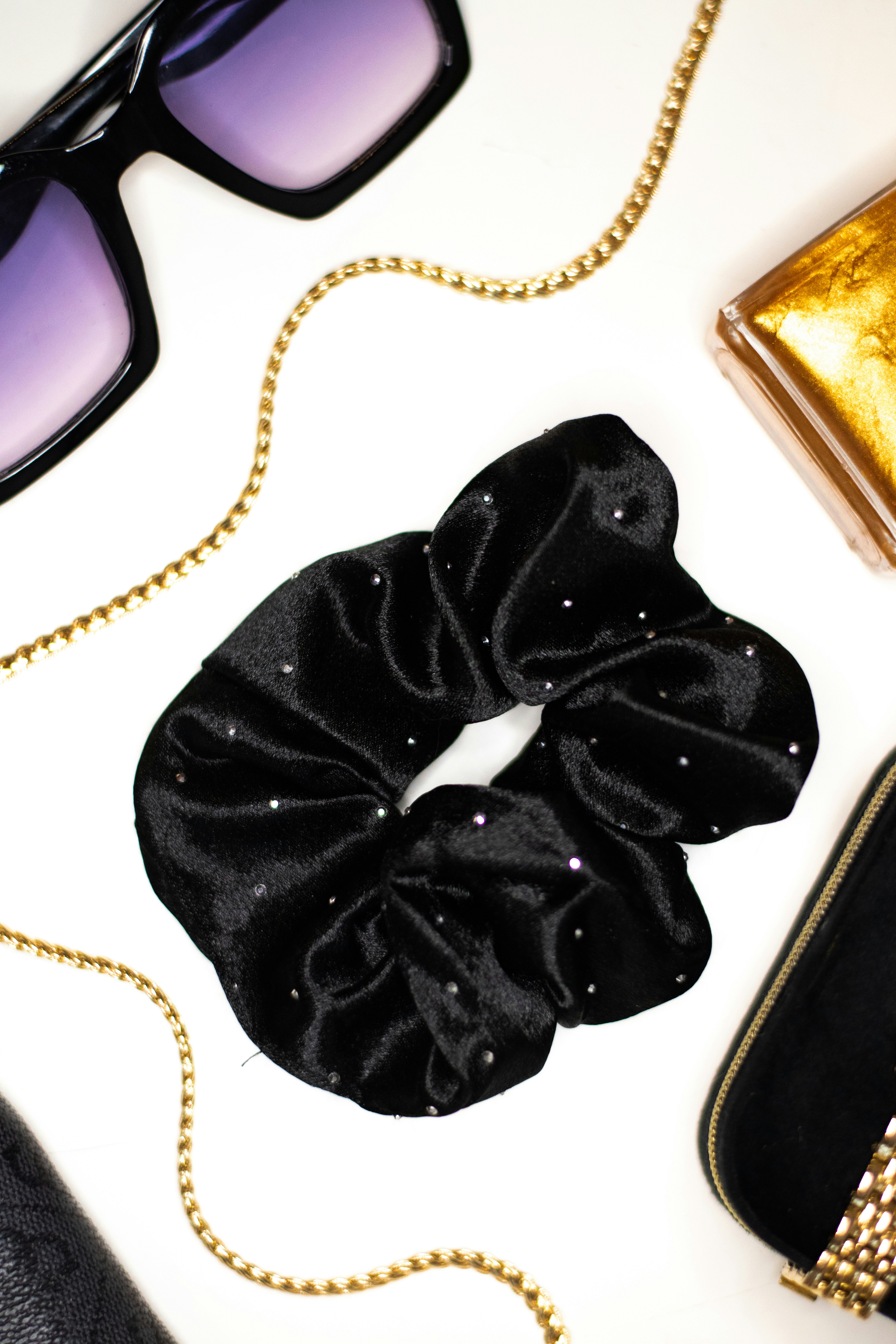 A black velvet scrunchie with small rhinestones surrounded by sunglasses, gold chain, and accessories