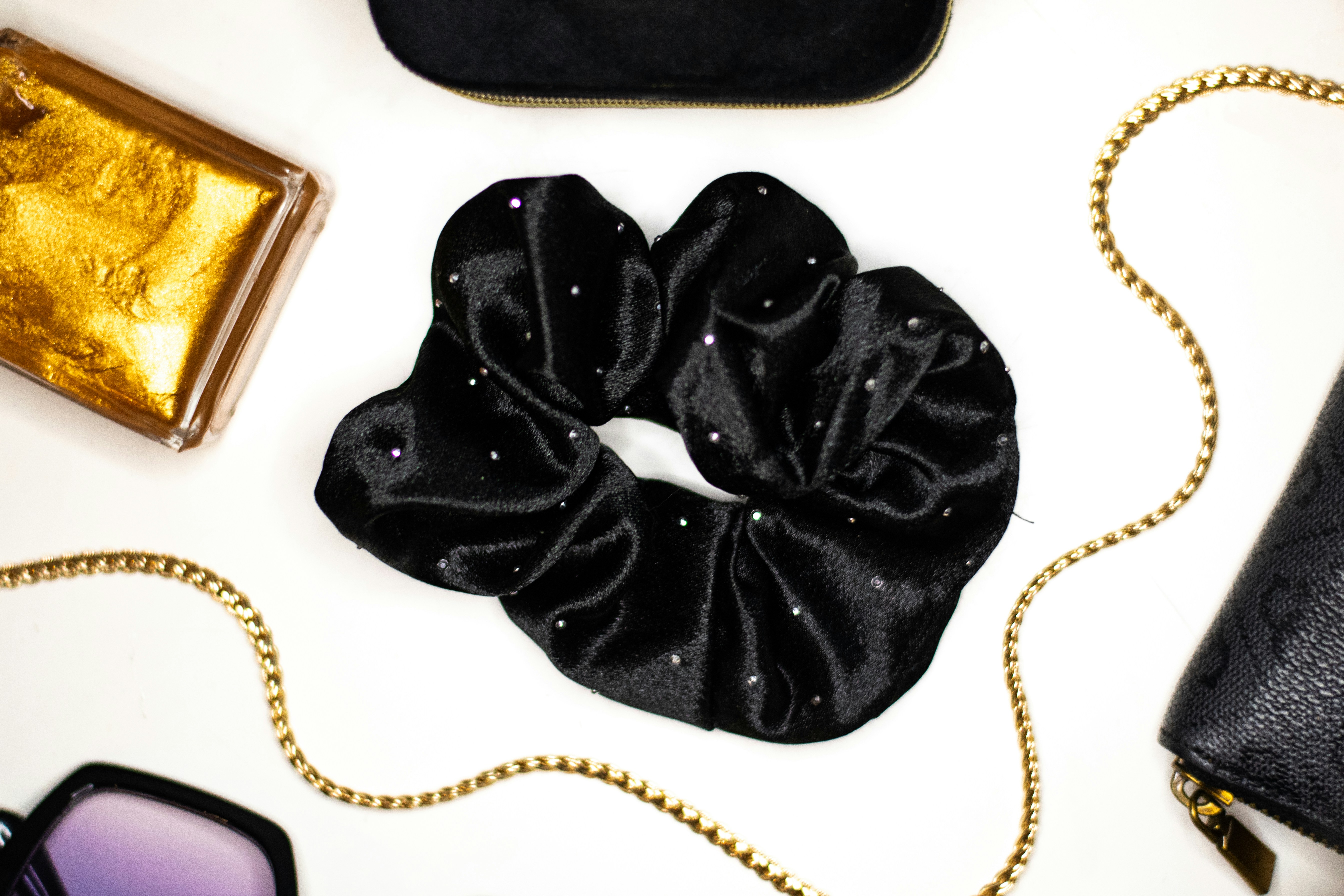 A black velvet scrunchie with small rhinestones beside gold accessories on a white surface