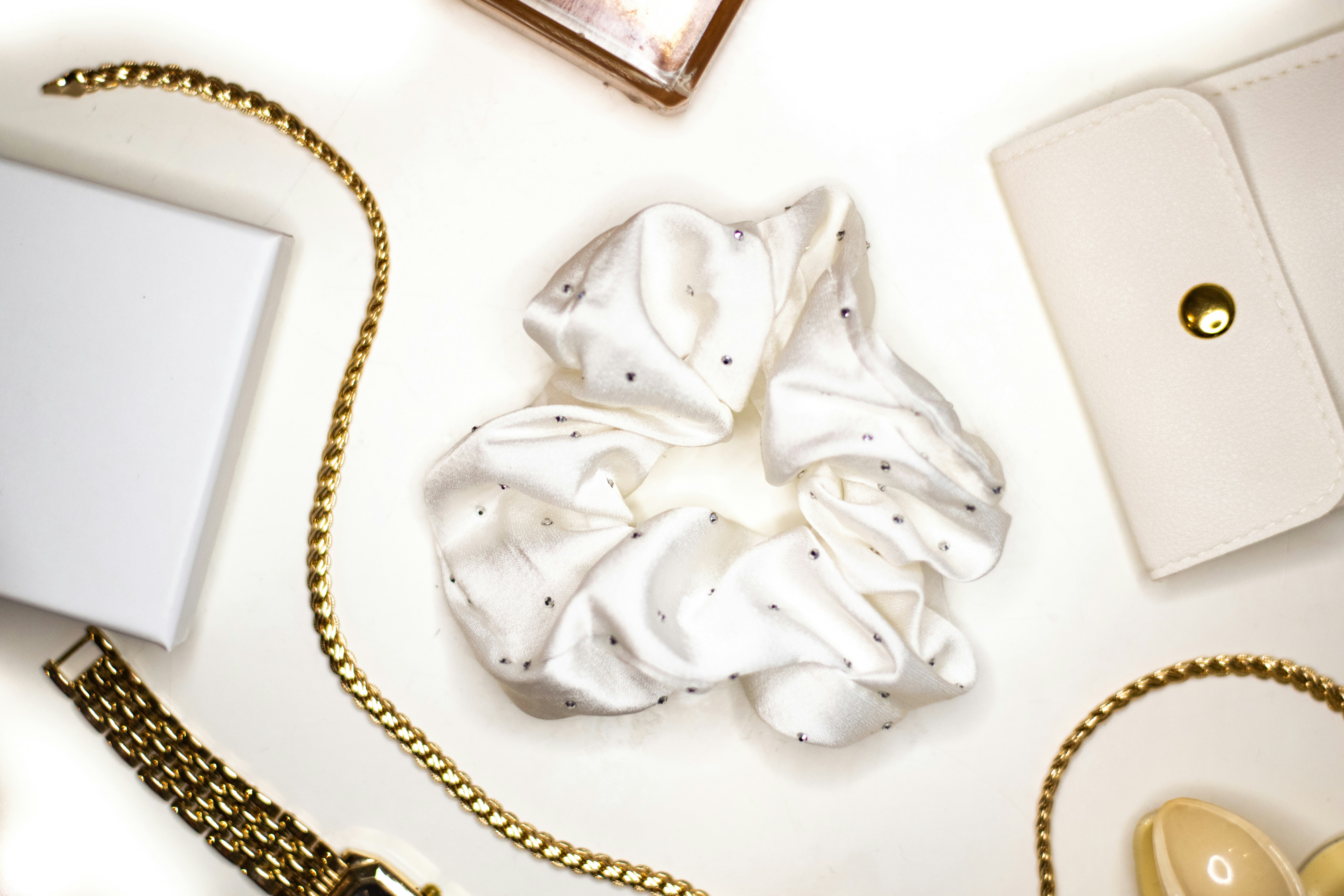 A white satin scrunchie with small crystals surrounded by gold jewelry and accessories