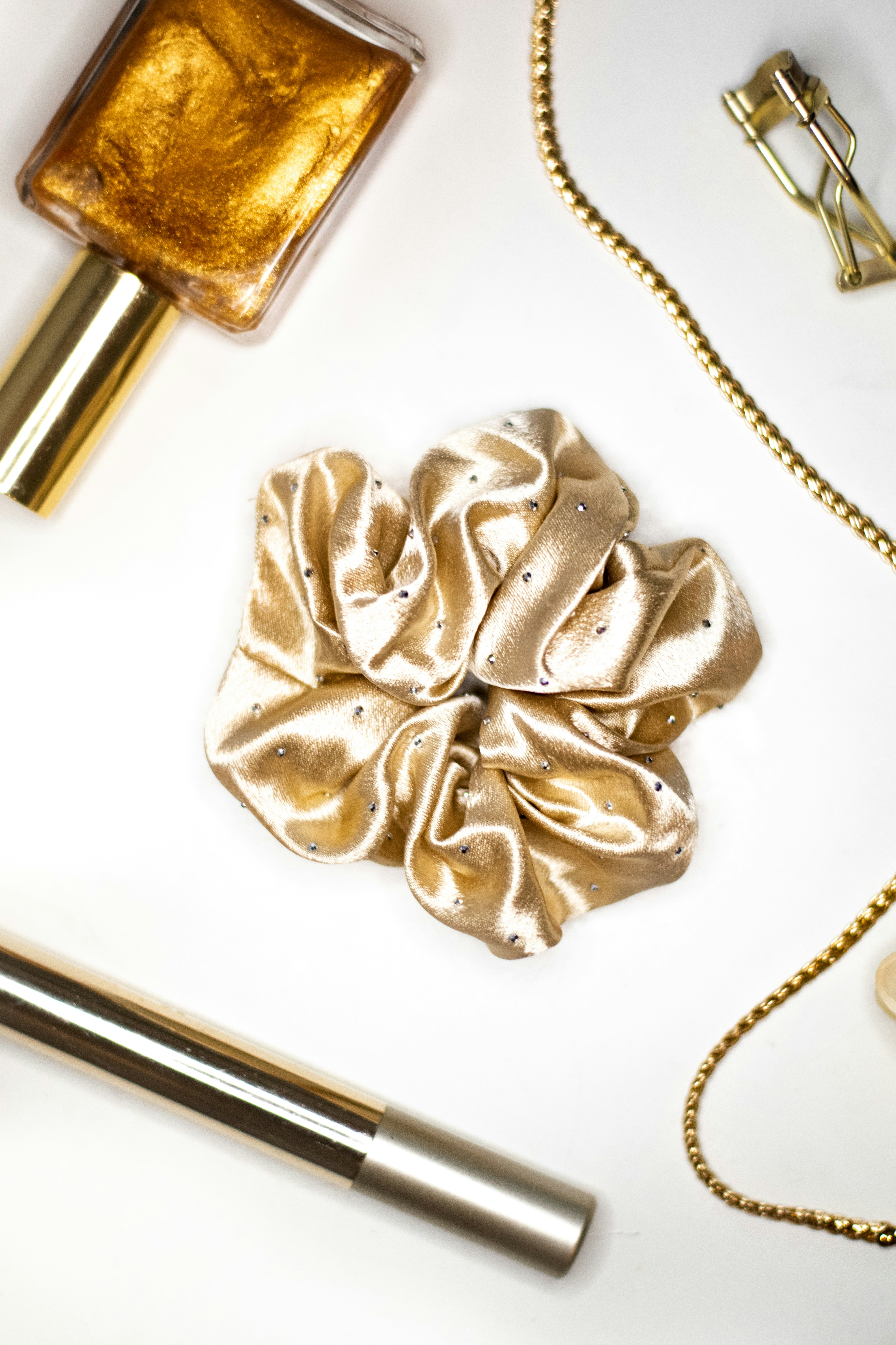 A gold satin scrunchie with small rhinestones beside nail polish and a gold chain