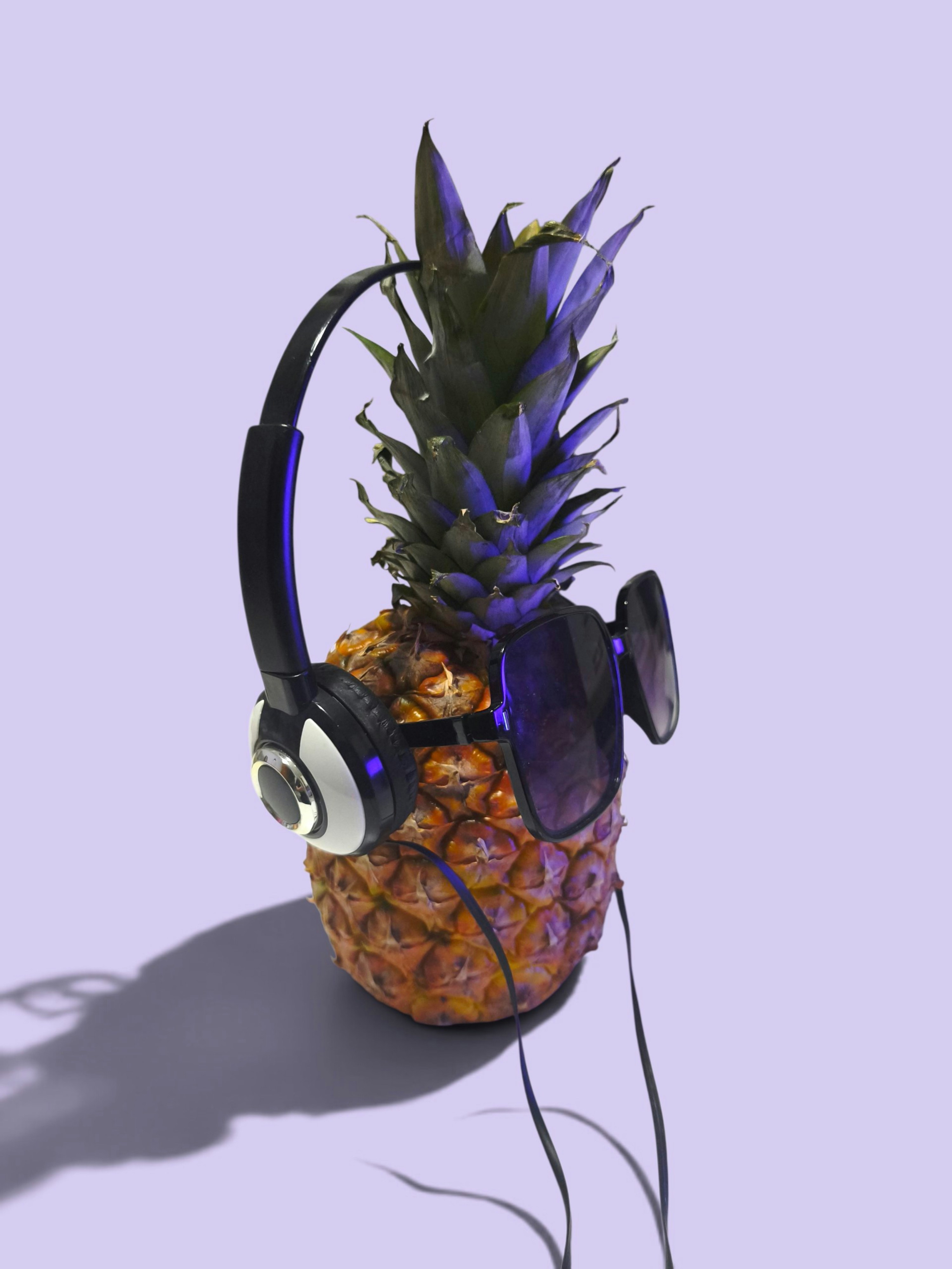 A pineapple wearing black headphones and sunglasses against a light purple background